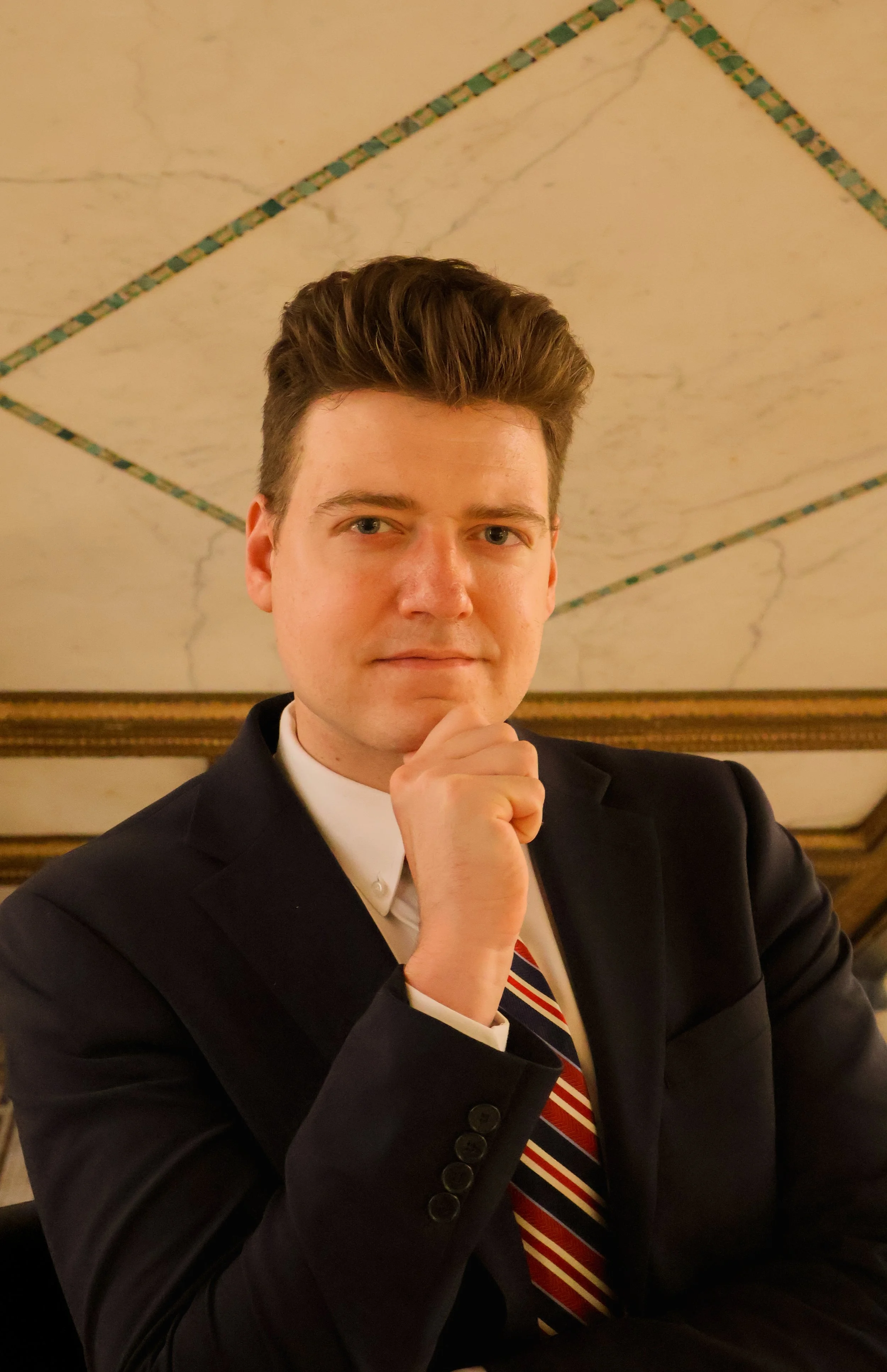 A man with brown hair and blue eyes wearing a navy suit, white shirt, and red striped tie, standing against a plain light-colored background.