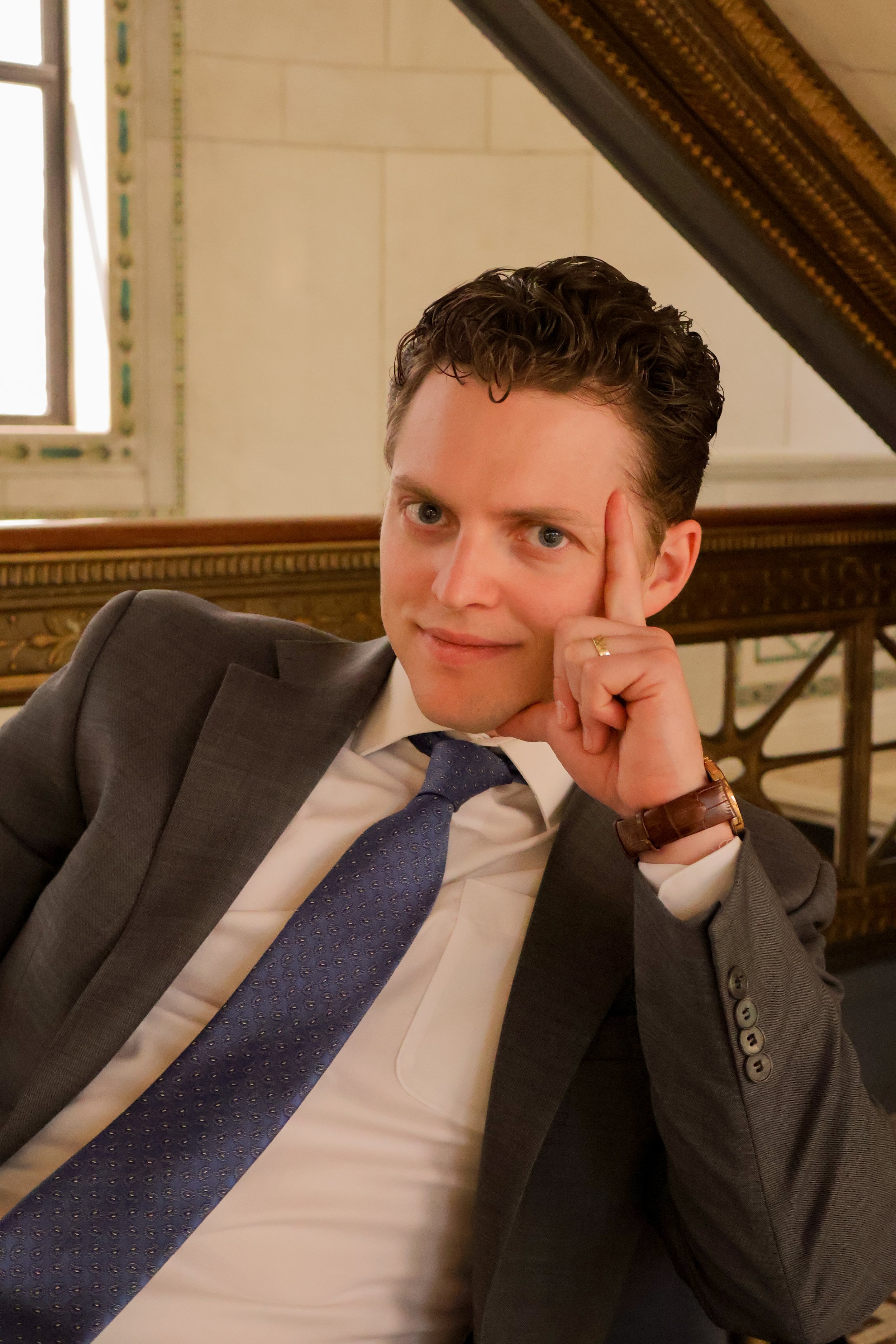 A young man with short, curly brown hair and blue eyes, wearing a dark navy suit, white dress shirt, and a blue patterned tie, standing against a plain light beige background.