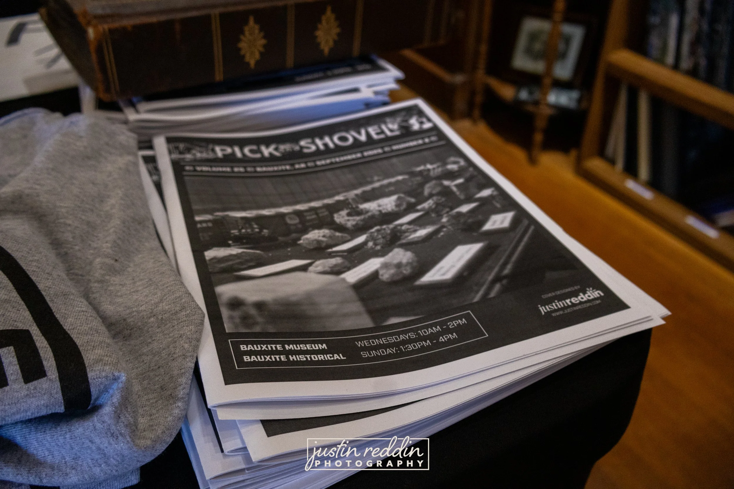 A stack of black and white posters advertising a rock climbing event at the Bauxite Museum with scheduled times on Wednesdays and Sundays. The posters are placed on a table next to a gray fabric item and a wooden bookcase.