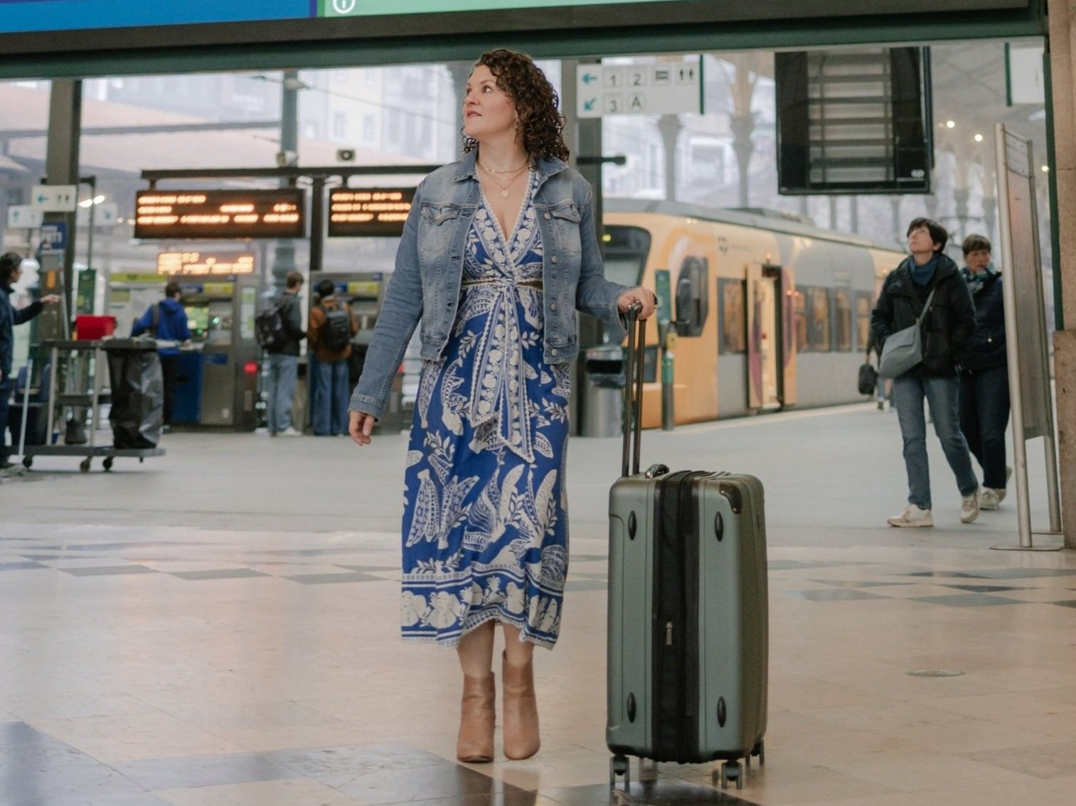 A woman in a blue and white patterned dress, tan boots, and a denim jacket walking with a rolling suitcase at a train station, with other travelers, a train, and station signs in the background.