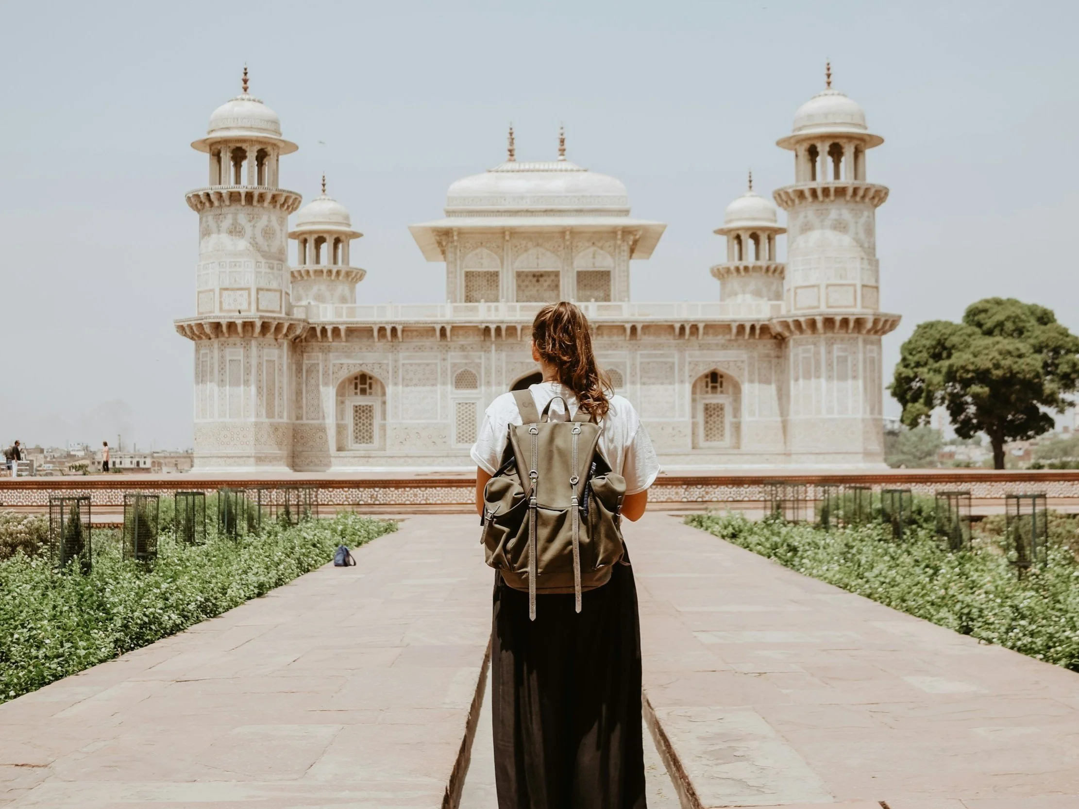 A woman with a backpack standing on a pathway, facing the Taj Mahal.