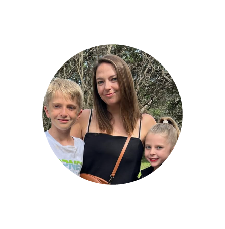 A woman with two children outdoors, smiling with trees in the background.