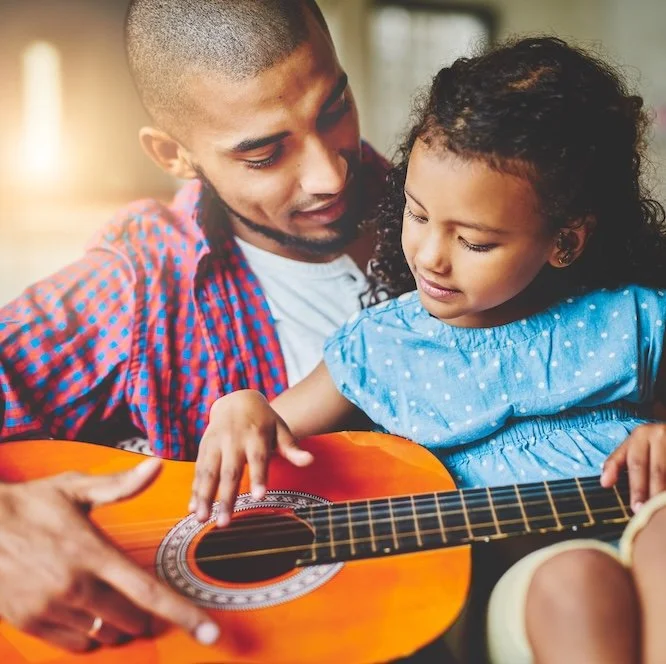 A young girl learning to play guitar with her father in a cozy, sunlit room.