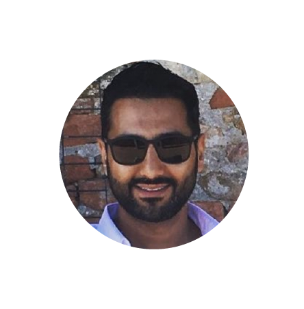 A man with dark hair, beard, and sunglasses smiling outdoors in front of a brick wall.