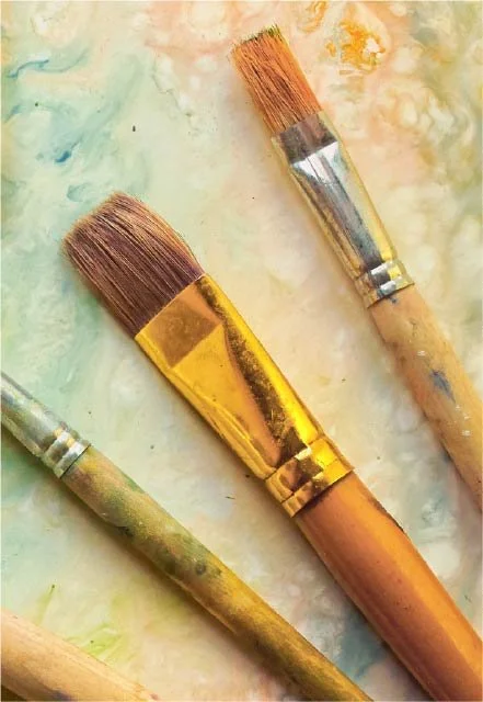 Three paintbrushes with wooden handles and bristles, resting on a colorful, painted surface.