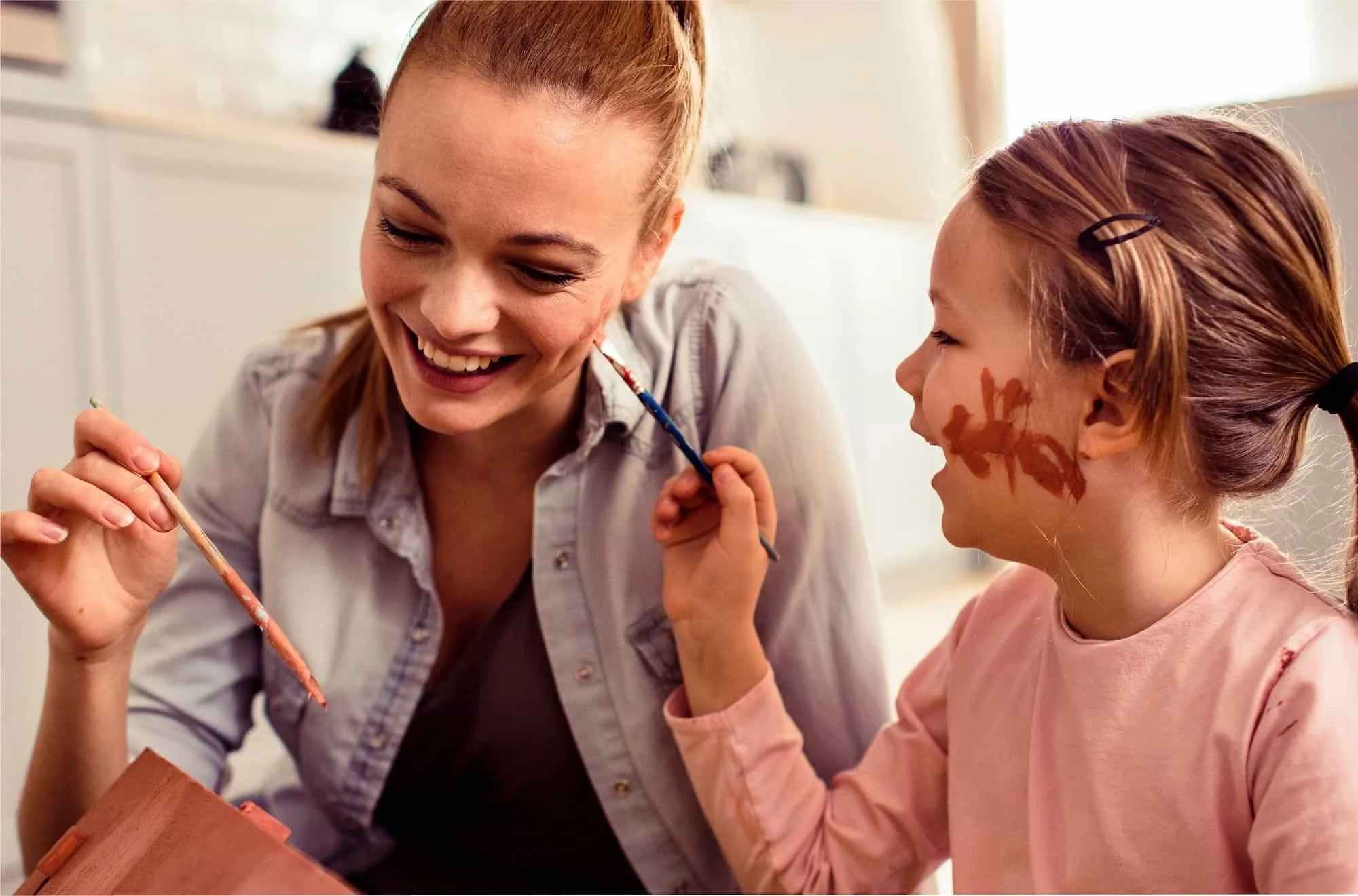 A woman and a young girl with face paint smiling and painting each other's faces indoors.