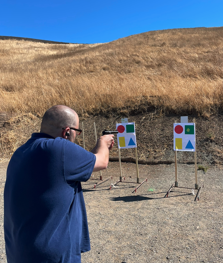 Man wearing headphones aiming a handgun at paper targets with geometric shapes in an outdoor shooting range.
