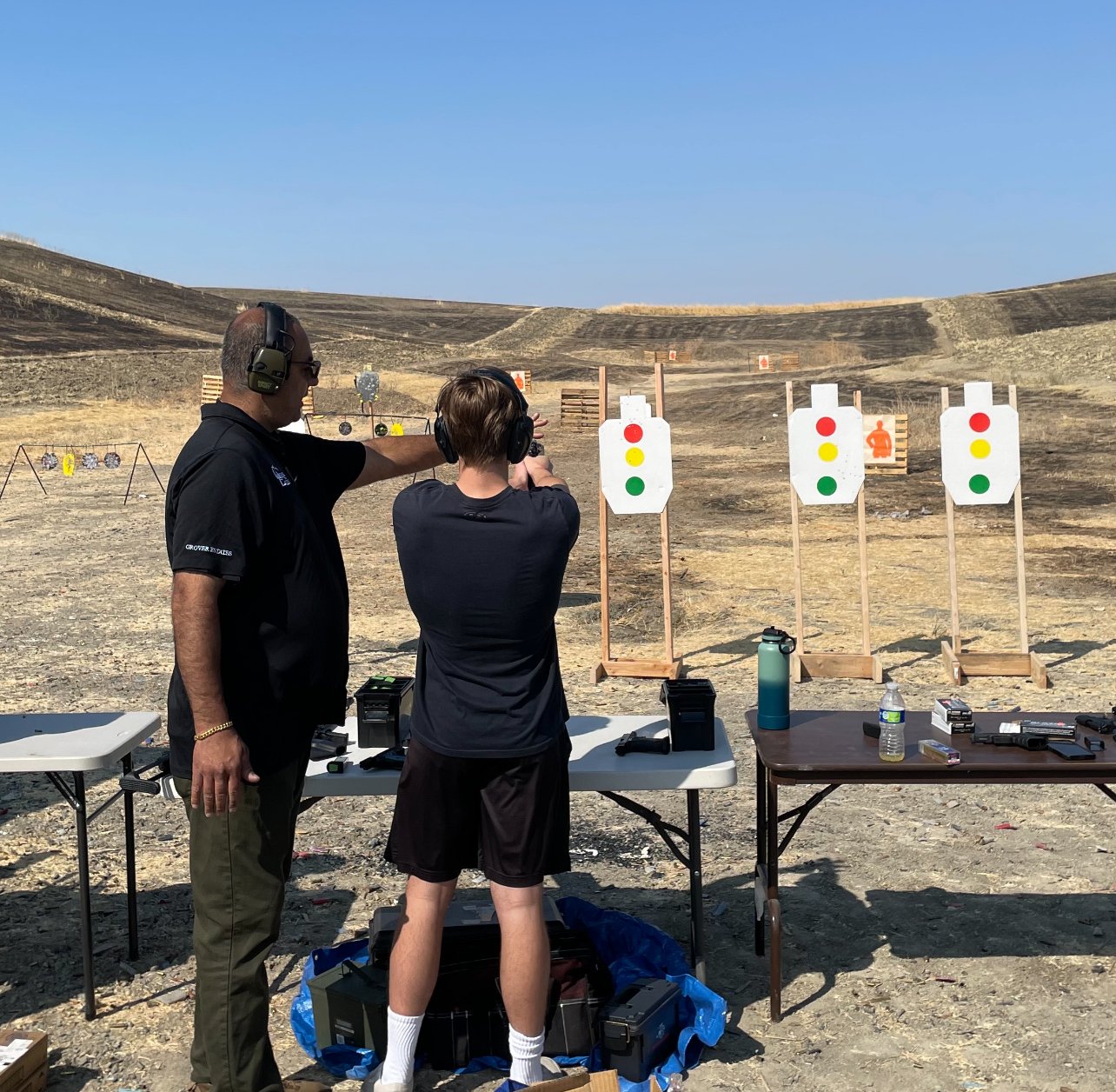 Two people at an outdoor shooting range, one instructing the other on aiming a handgun at paper targets with traffic light symbols. They are wearing hearing protection, and there are tables with equipment nearby.
