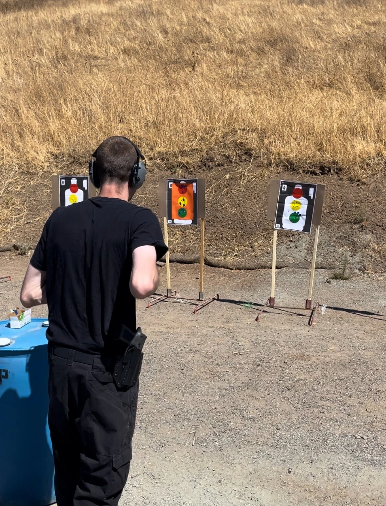 Person at shooting range aiming at paper targets with ear protection and holstered firearm.