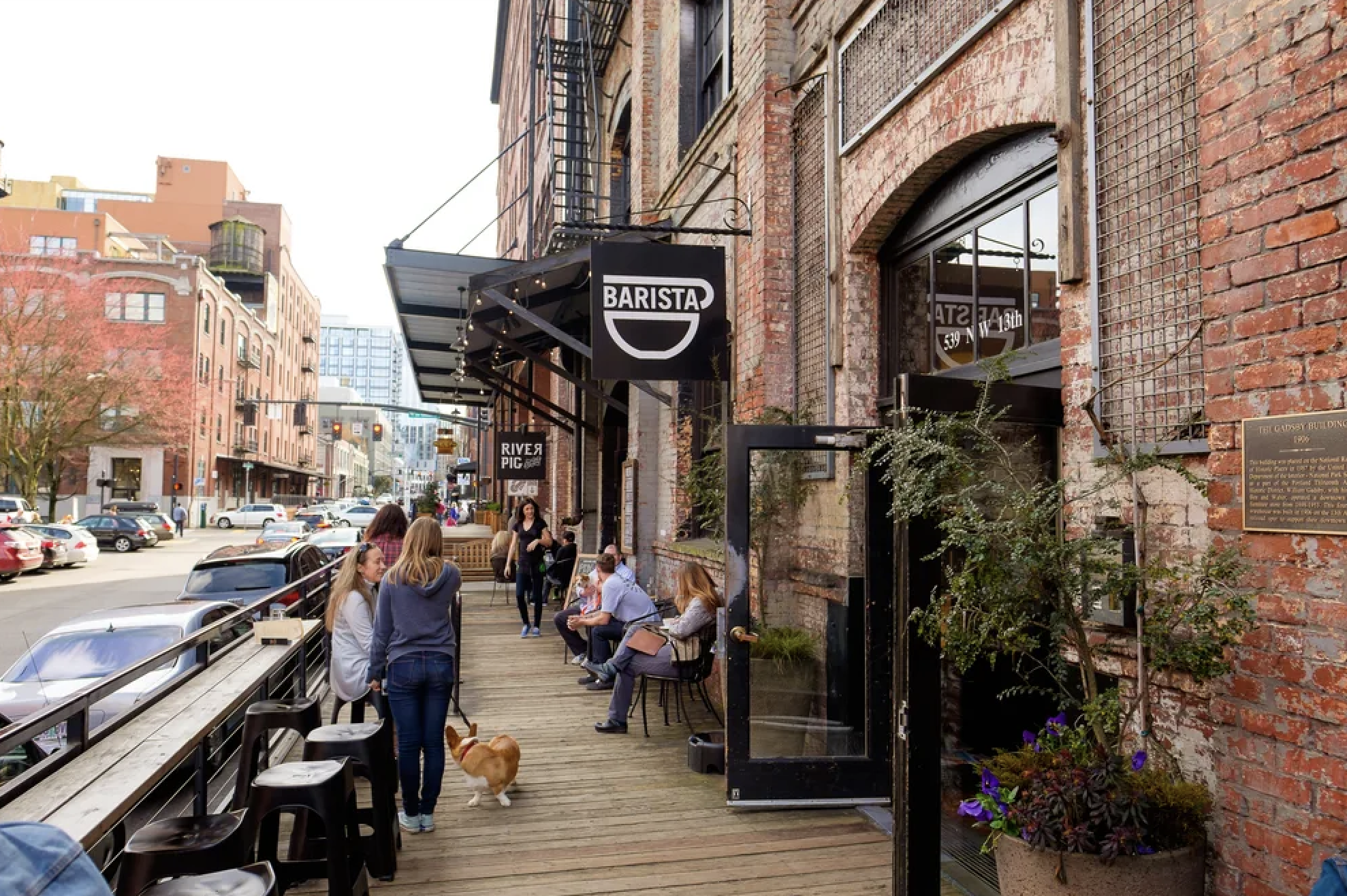 People sitting and walking on a wooden sidewalk outside a brick building with signs reading 'Barista' and 'River Pig'. A woman is walking her dog, and others are seated at outdoor tables, with trees and parked cars nearby.