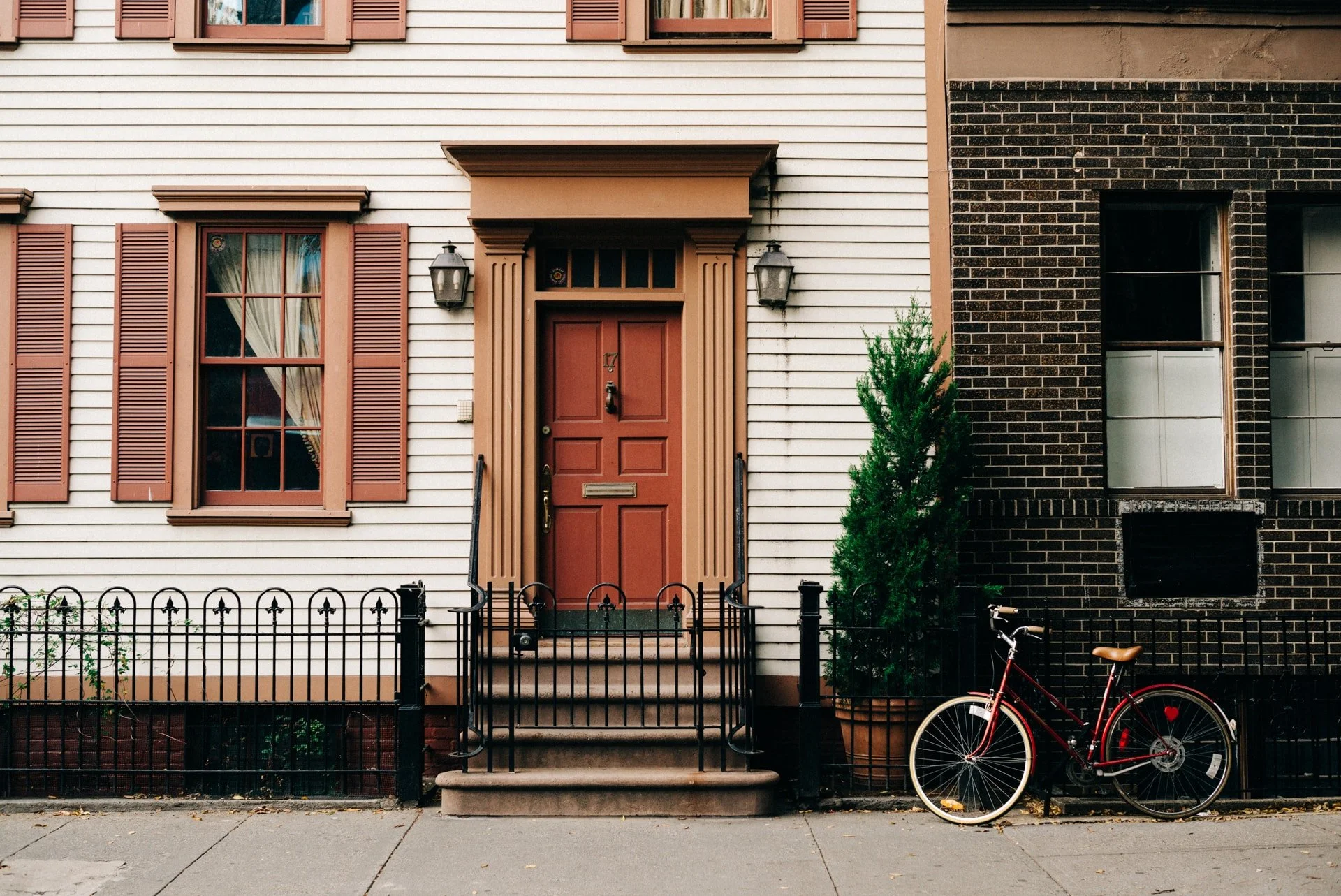 A house with white and black brick exterior walls, a red front door, and a red bicycle parked outside. The house has brown shutters and lantern-style lights beside the front door. A small green shrub is to the right of the door.