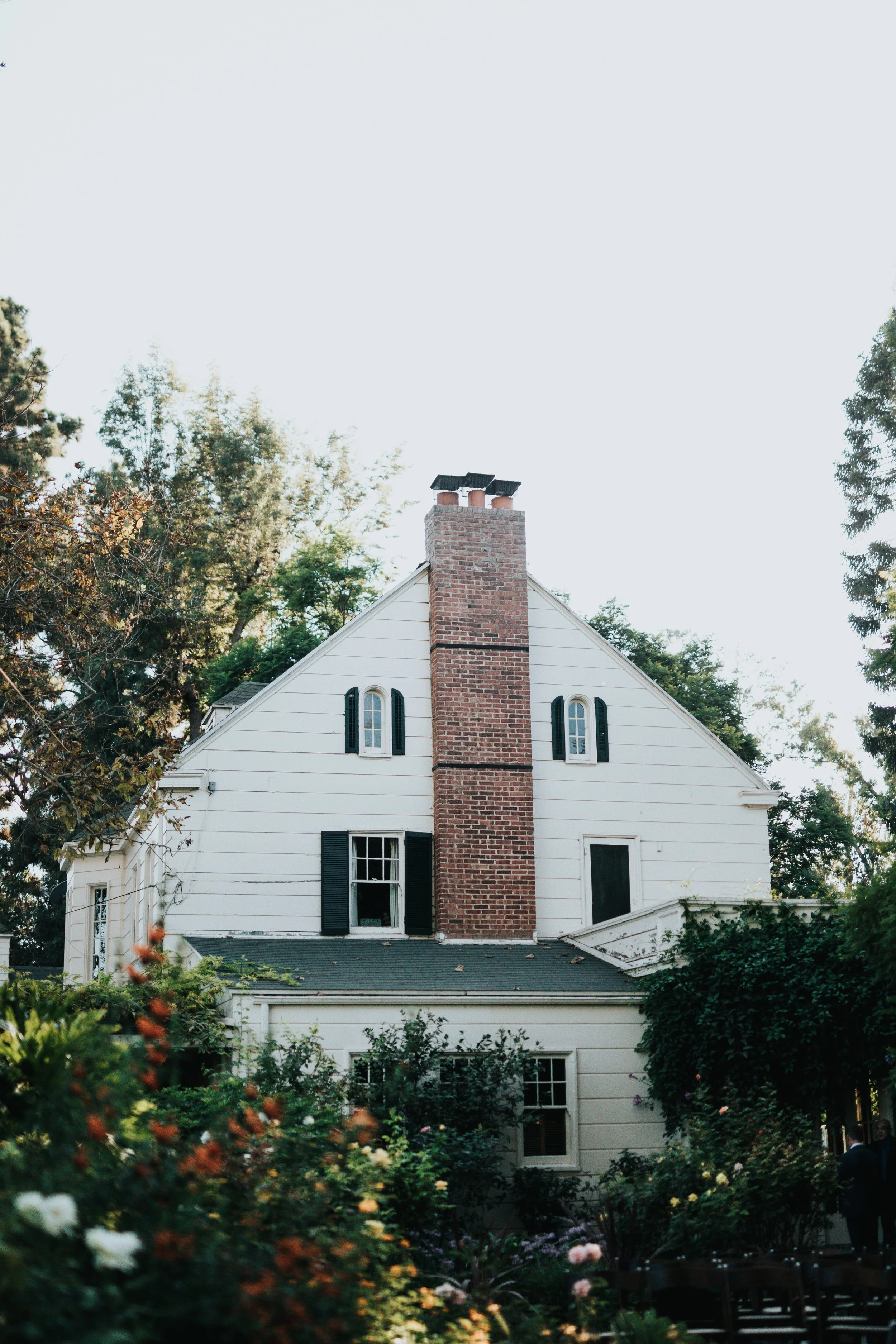 A white house with a brick chimney, surrounded by trees and a garden with flowers.