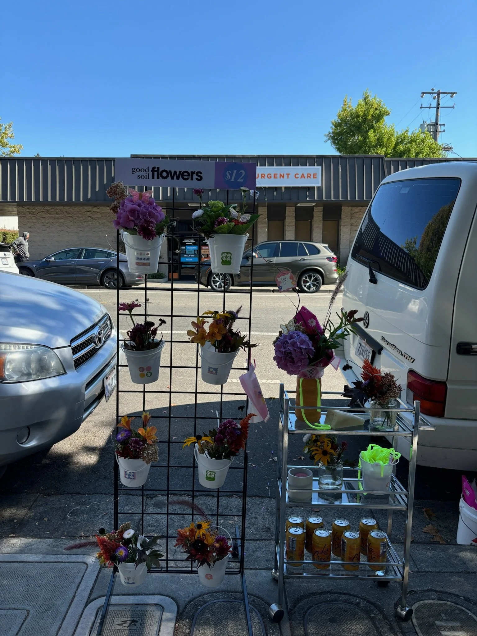 Flower stand with various colorful bouquets in white buckets and a metal cart with additional flowers and canned drinks on a sidewalk in front of a parking lot and a storefront sign that reads 'good soil flowers' and 'urgent care'.