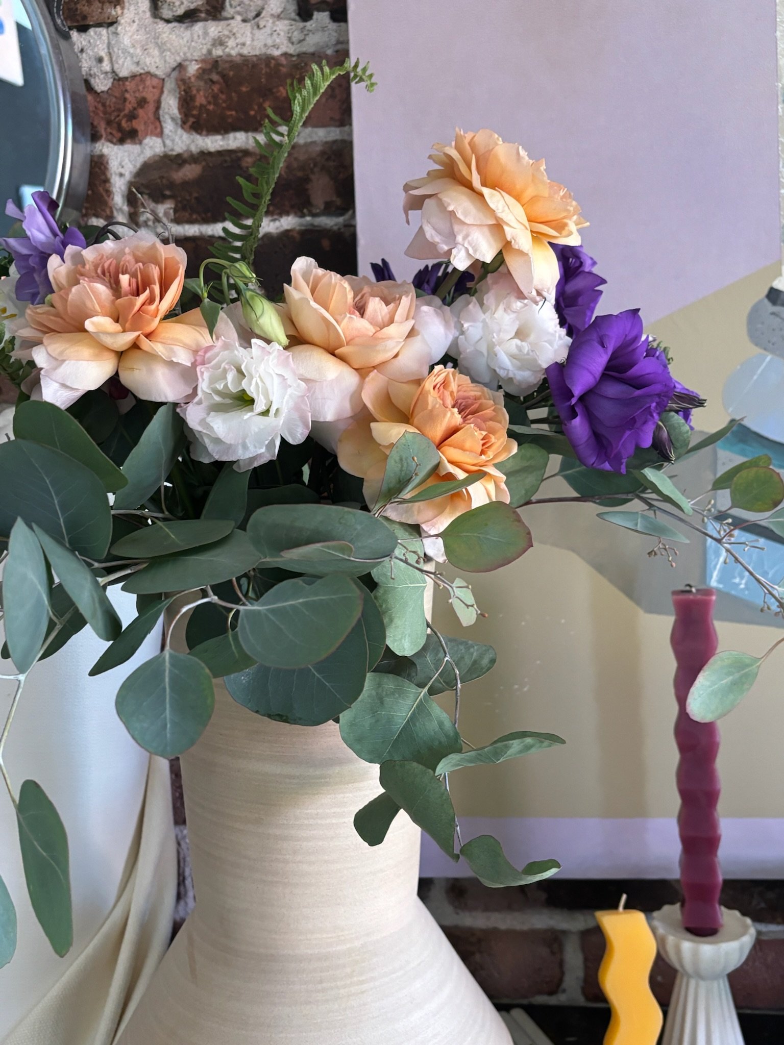A bouquet of flowers in a cream-colored vase, featuring peach, white, and purple blooms, with green eucalyptus leaves against a brick wall background.
