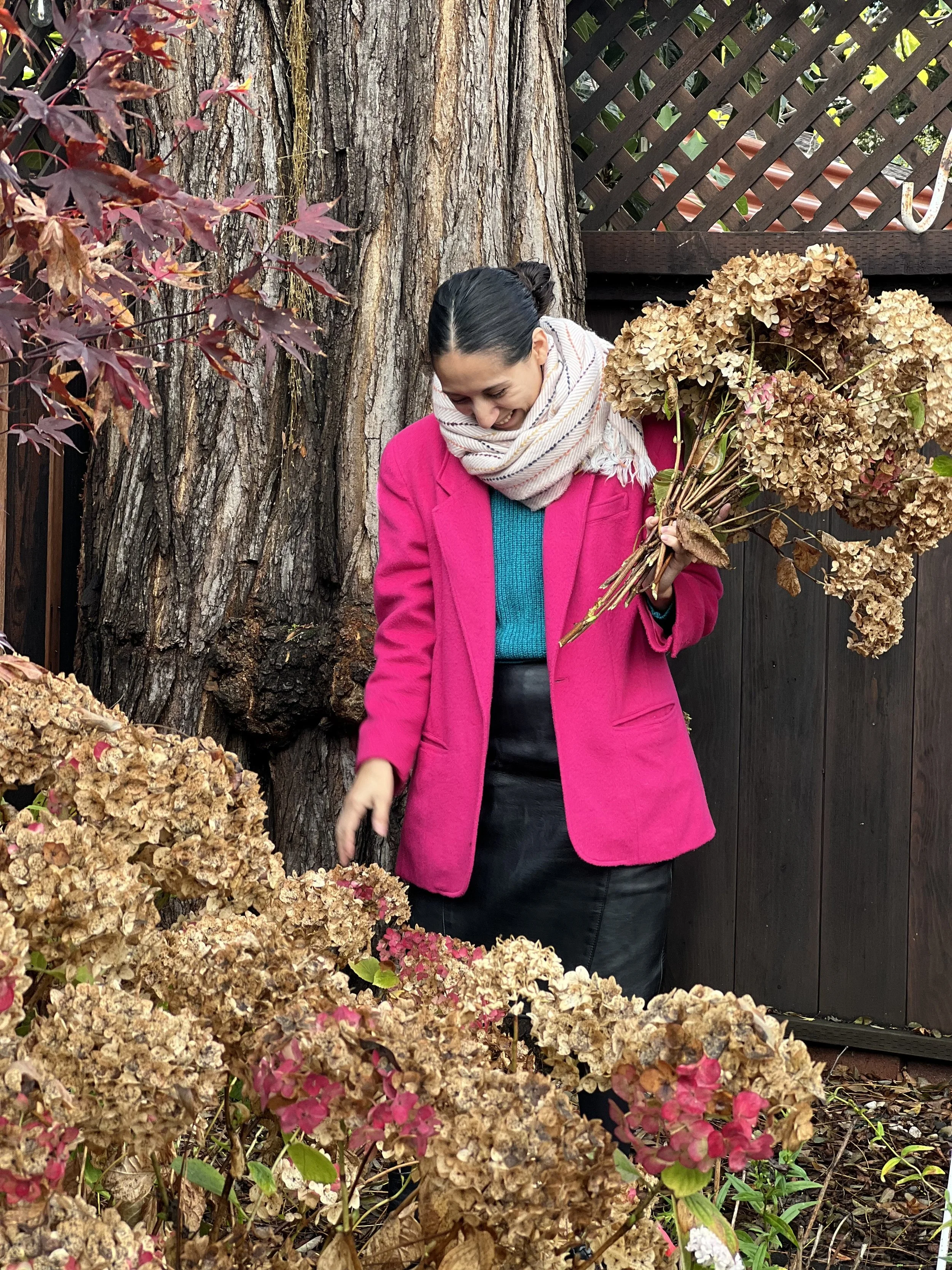 A woman wearing a bright pink coat, blue sweater, and scarf, holding a bunch of dried hydrangea flowers and smiling while standing outdoors near a large tree and a wooden fence.