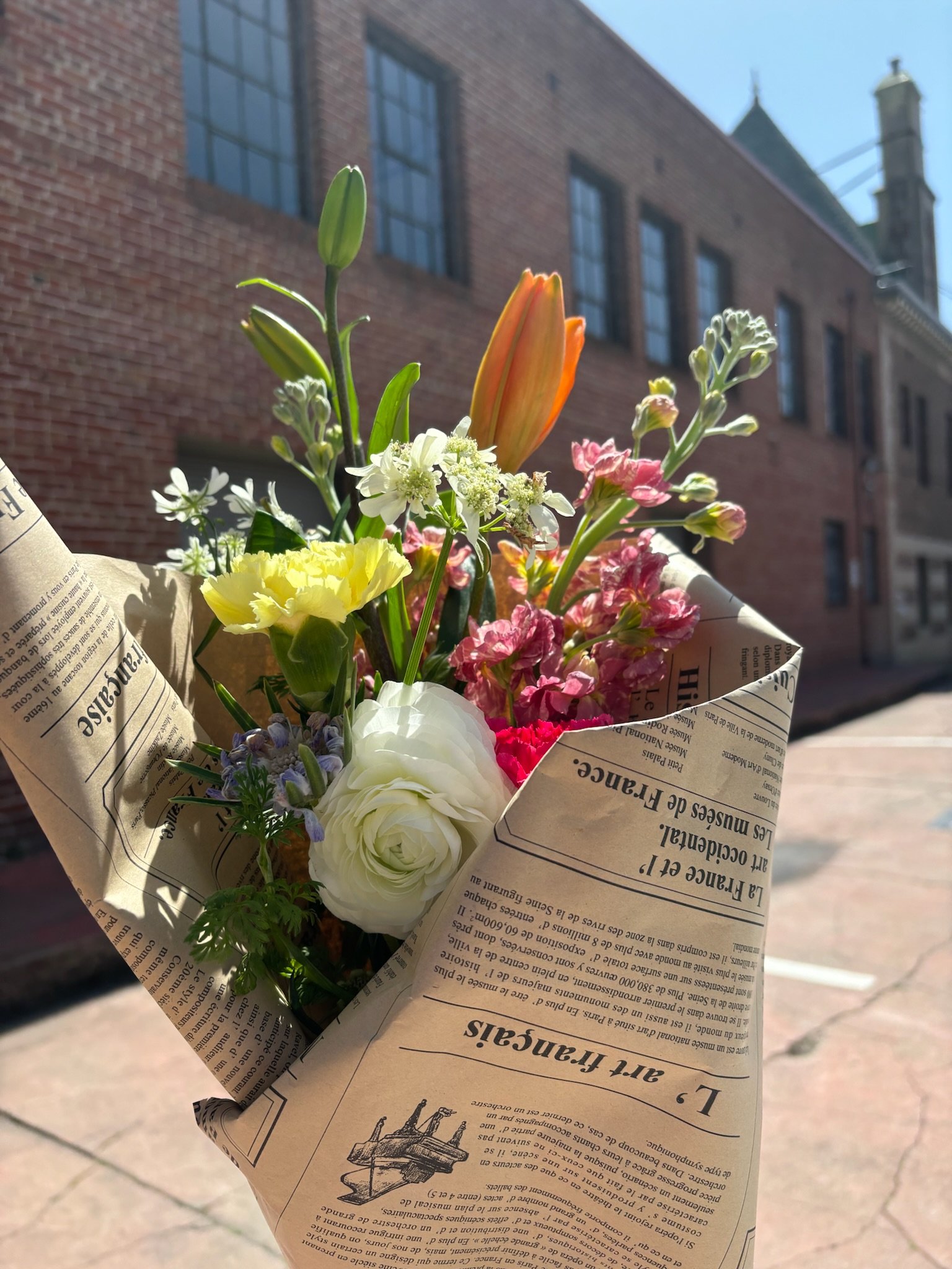 Bouquet of mixed flowers wrapped in newspaper, held outdoors on a sunny day.