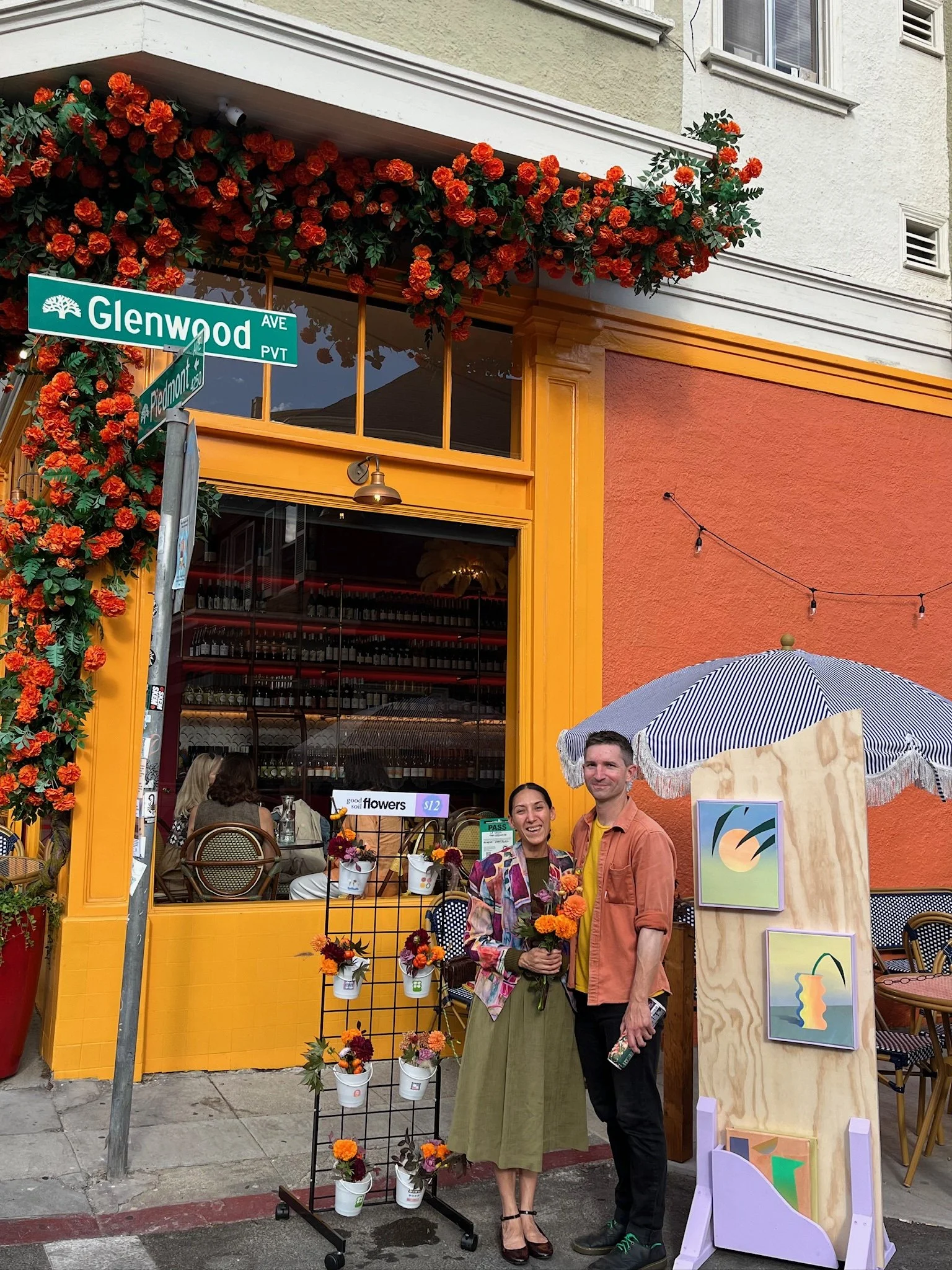 A couple posing outside a vibrant orange and yellow storefront on a city corner. The woman holds a bouquet of flowers and is smiling, wearing a colorful jacket and a long green skirt. The man stands next to her, holding a drink, wearing a peach-color