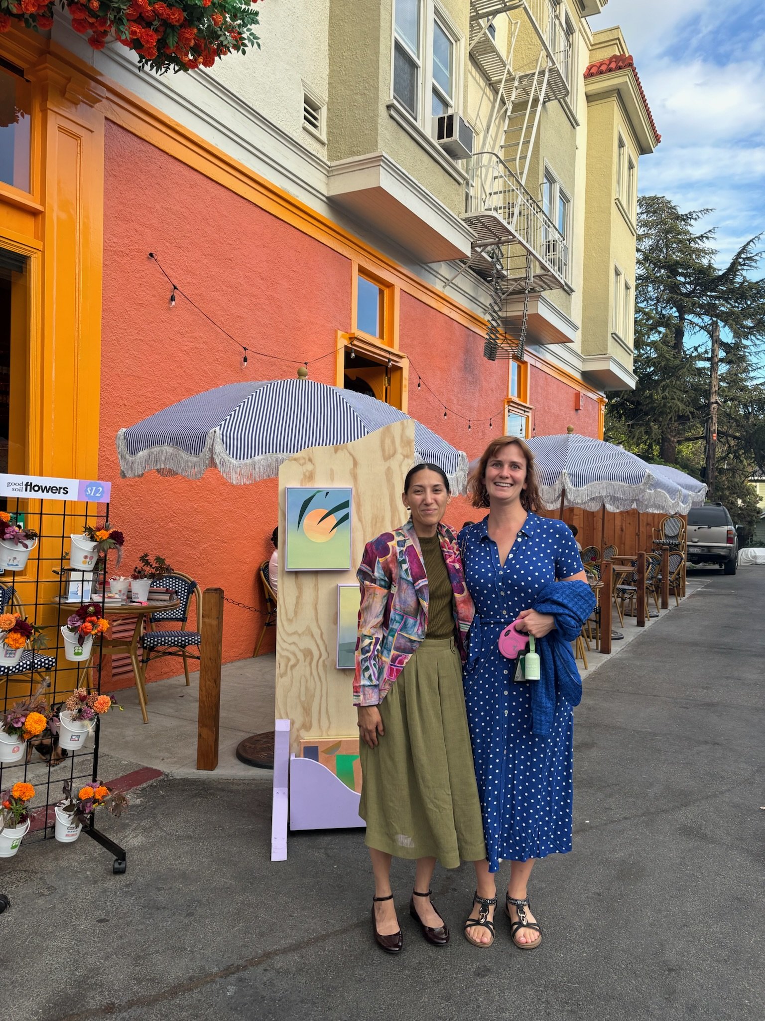 Two women standing outside a colorful building with pink and yellow walls, under patio umbrellas. One woman is wearing a patterned jacket and olive skirt, the other a blue polka dot dress, holding a pink purse and a sanitizer bottle.