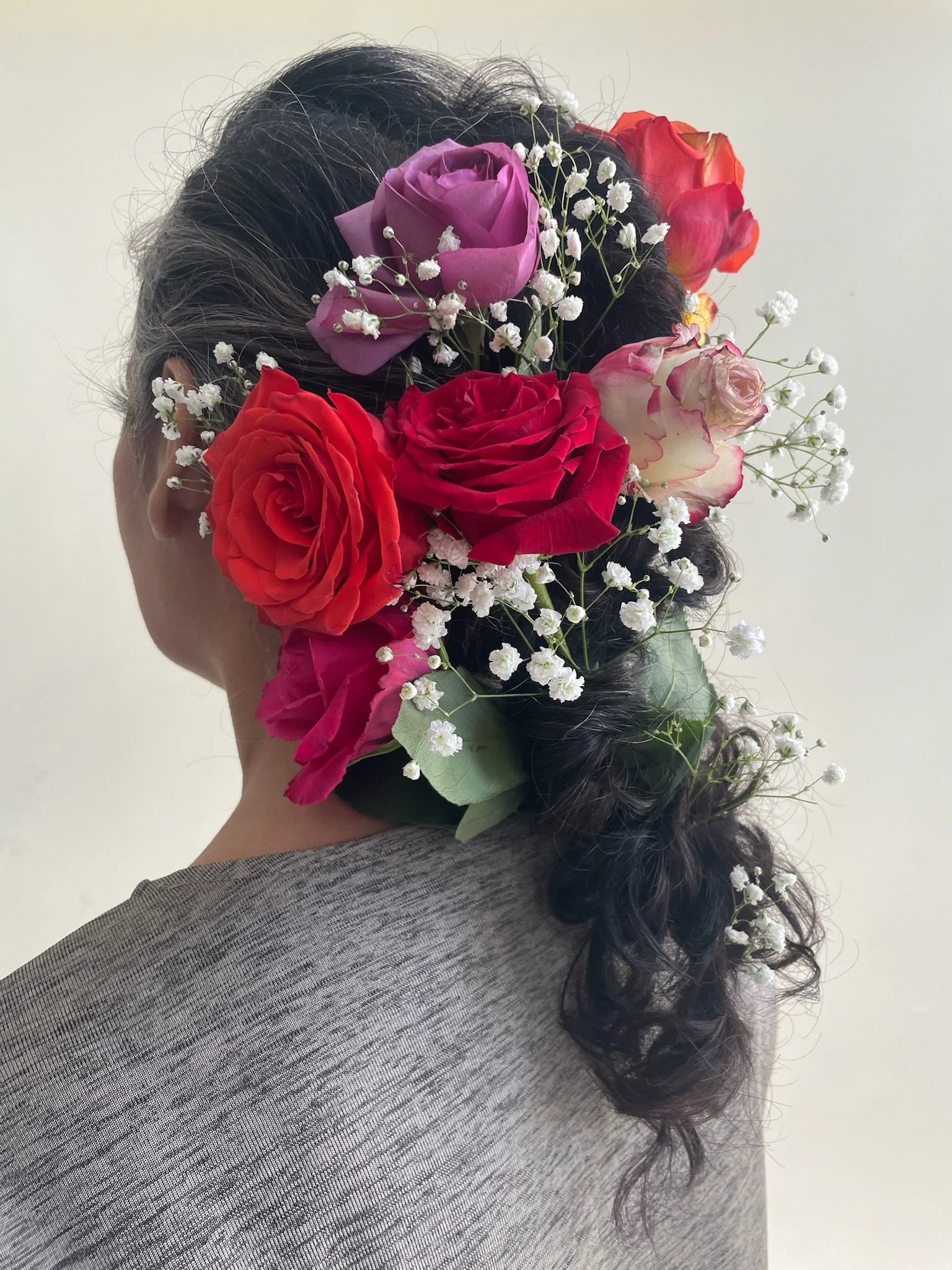 A woman with dark curly hair styled in a loose ponytail, wearing a gray top, has a floral arrangement covering the back of her head with pink roses, red roses, purple roses, baby's breath, and green leaves.