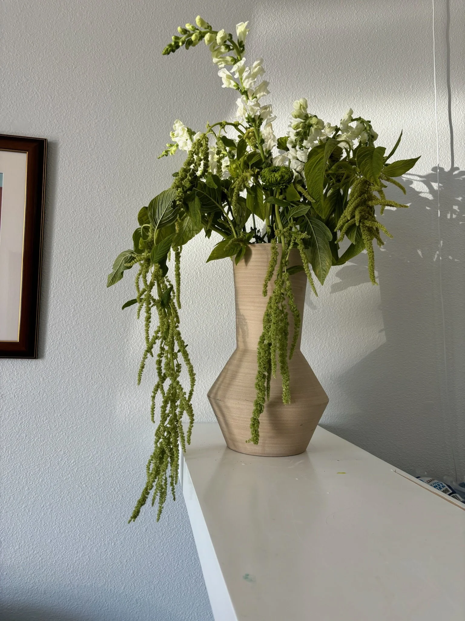 A beige, faceted ceramic vase on a white surface holding a bouquet of white snapdragons, green leaves, and trailing green amaranth plants against a light-colored wall.