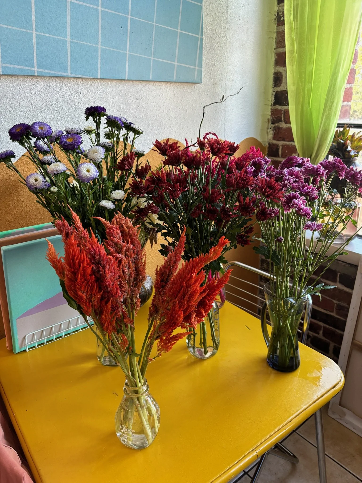 Vases of colorful flowers on a yellow table near a window with green curtains in a cozy room.