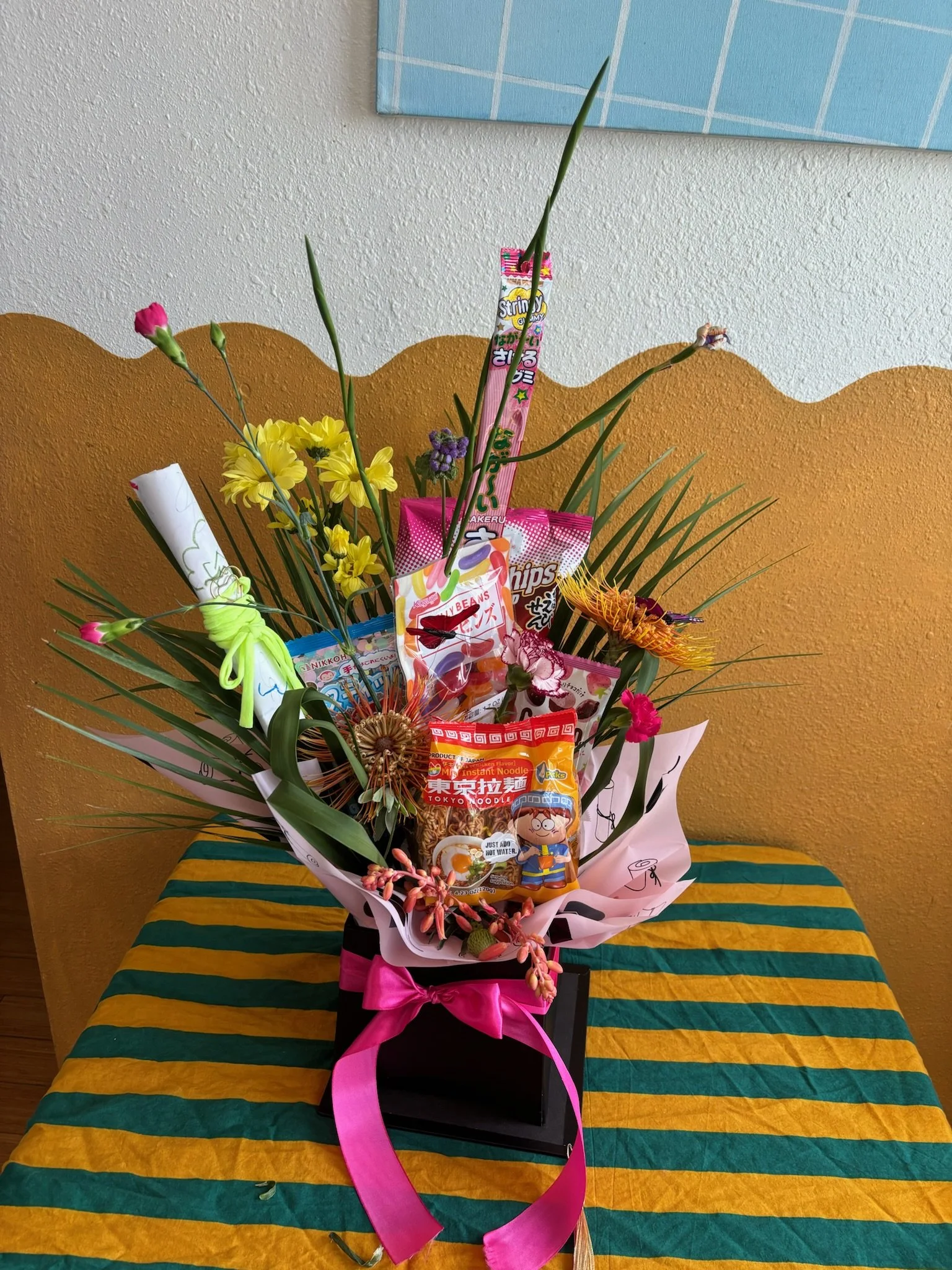 A colorful bouquet with flowers and Japanese snacks arranged in a black box with a pink ribbon, placed on a table with a striped green and yellow tablecloth.