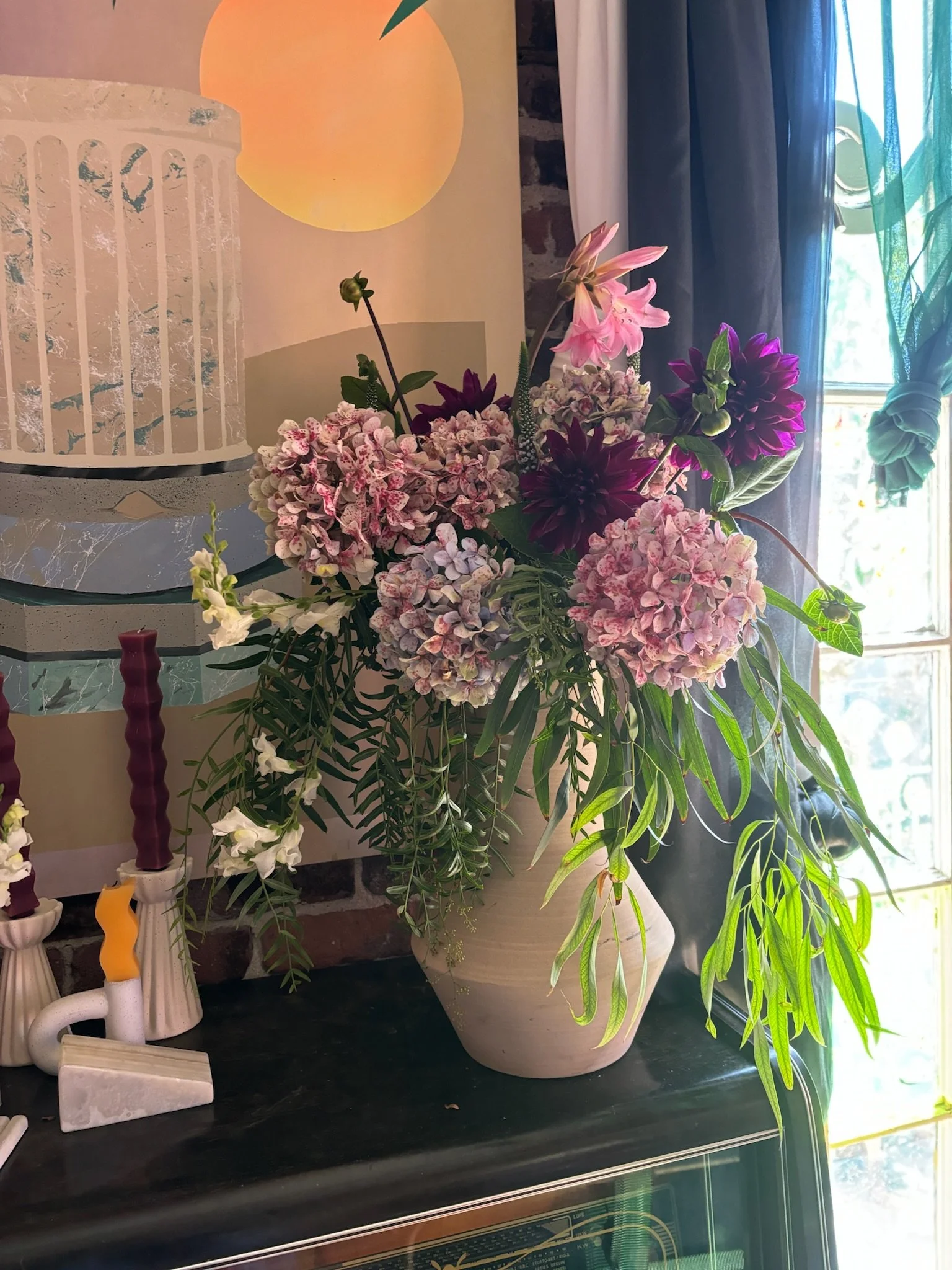 A bouquet of pink, purple, and white flowers arranged in a large off-white ceramic vase on a dark surface near a window with sunlight, curtains, and a brick wall, with a modern art print and some candles nearby.