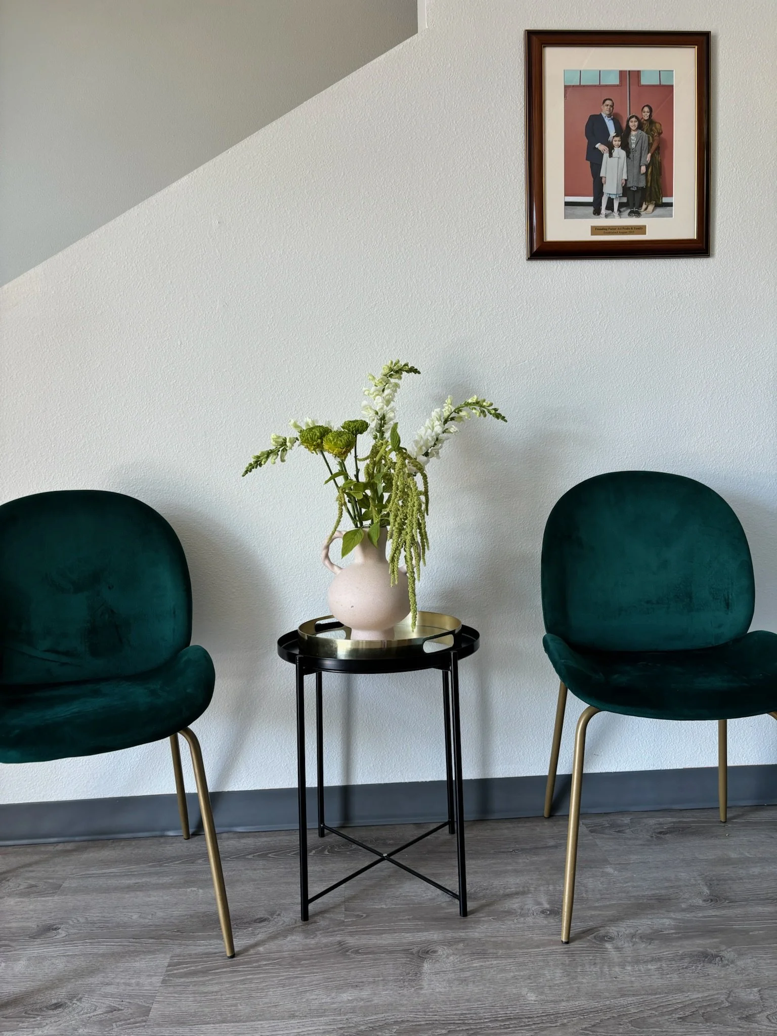 Two green velvet chairs on either side of a round black side table with a white vase containing white and green flowers, with a framed family photo hanging on a white wall above.