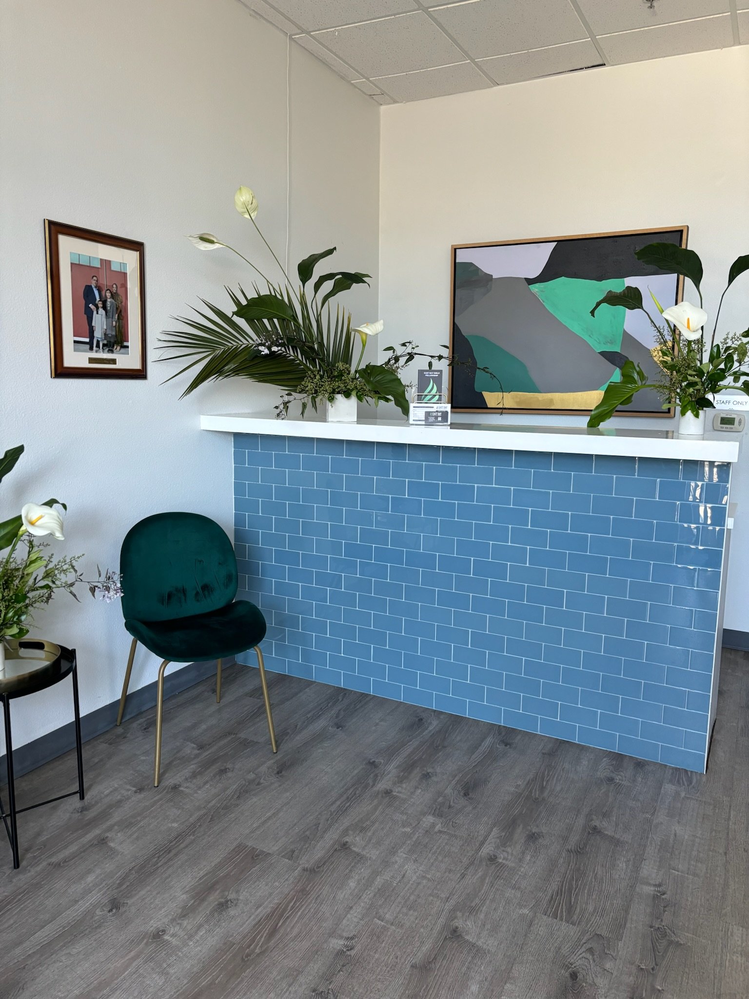 Reception area with a teal brick counter, green velvet chair, plants, and framed artwork on white walls.