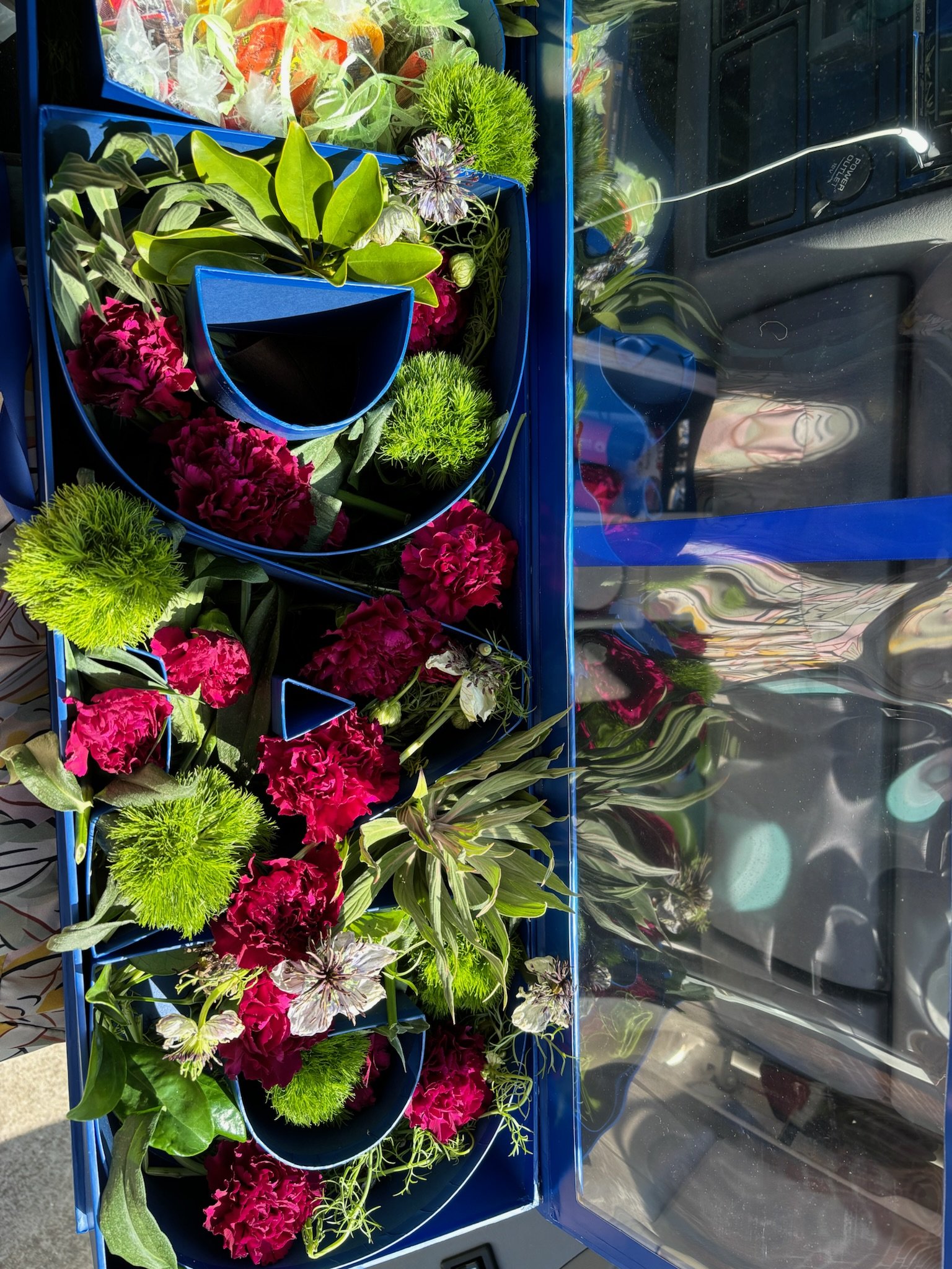 A blue tray with various colorful flowers, including pink carnations, green pom-pom flowers, and variegated leaves, inside a vehicle with the door open.