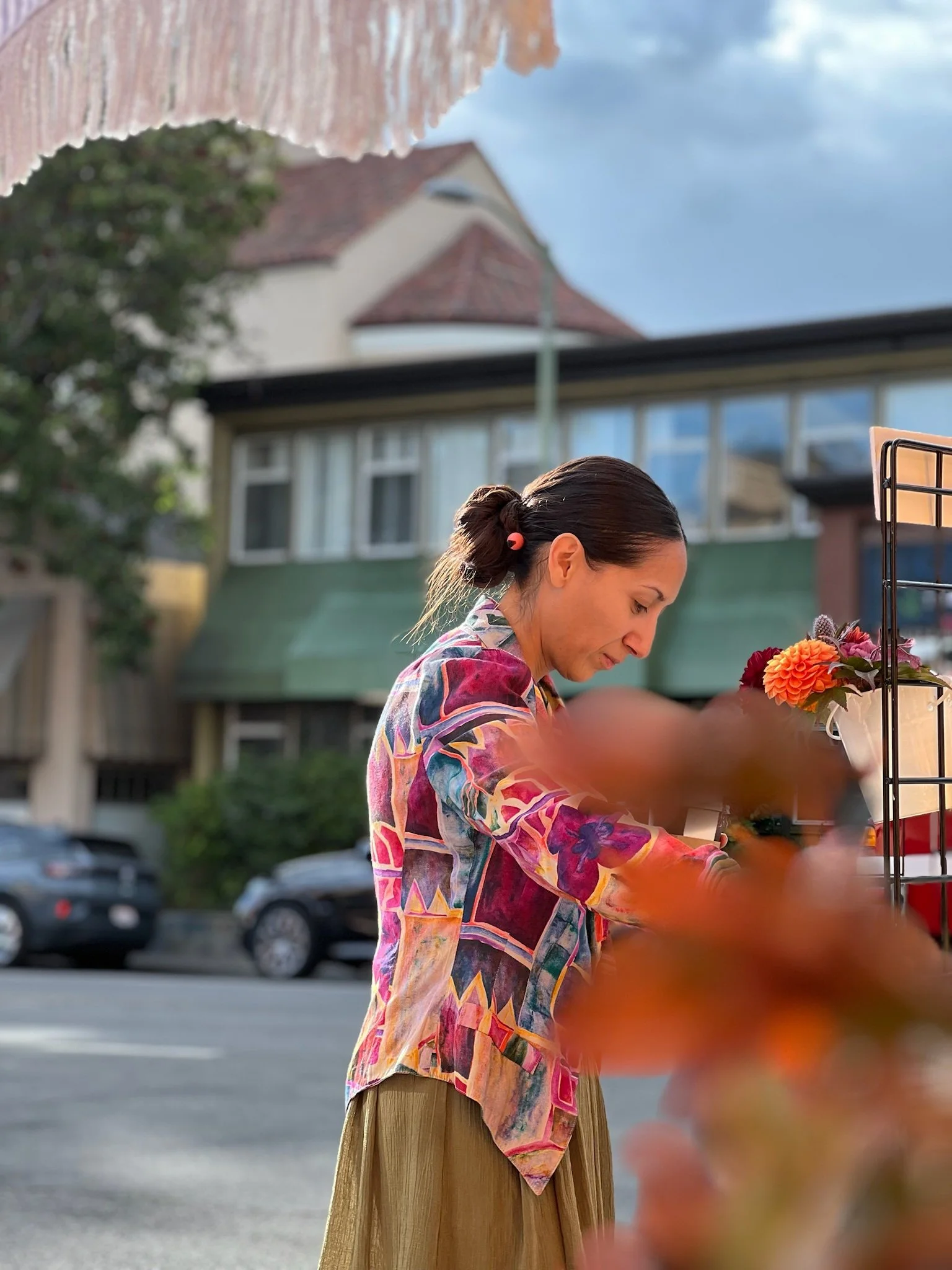 A woman with dark hair tied back, wearing a colorful, patterned blouse and a beige skirt, standing on a street outdoors. She appears to be looking at or arranging flowers on a display stand. Cars are parked in the background, and there are buildings 