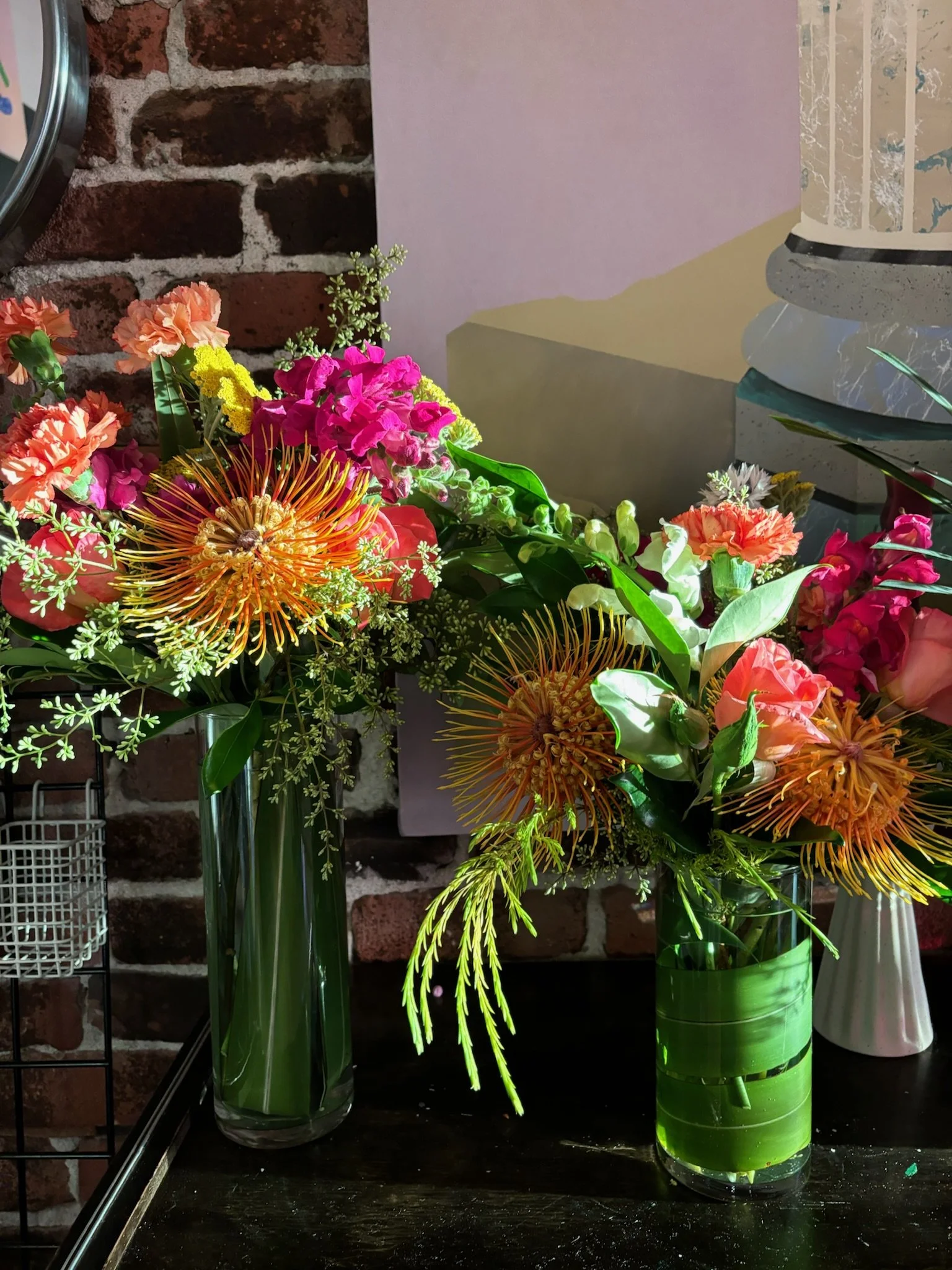Two glass vases with colorful flowers, including orange pincushion proteas, pink and peach carnations, and greenery, placed on a dark surface against a brick wall background.