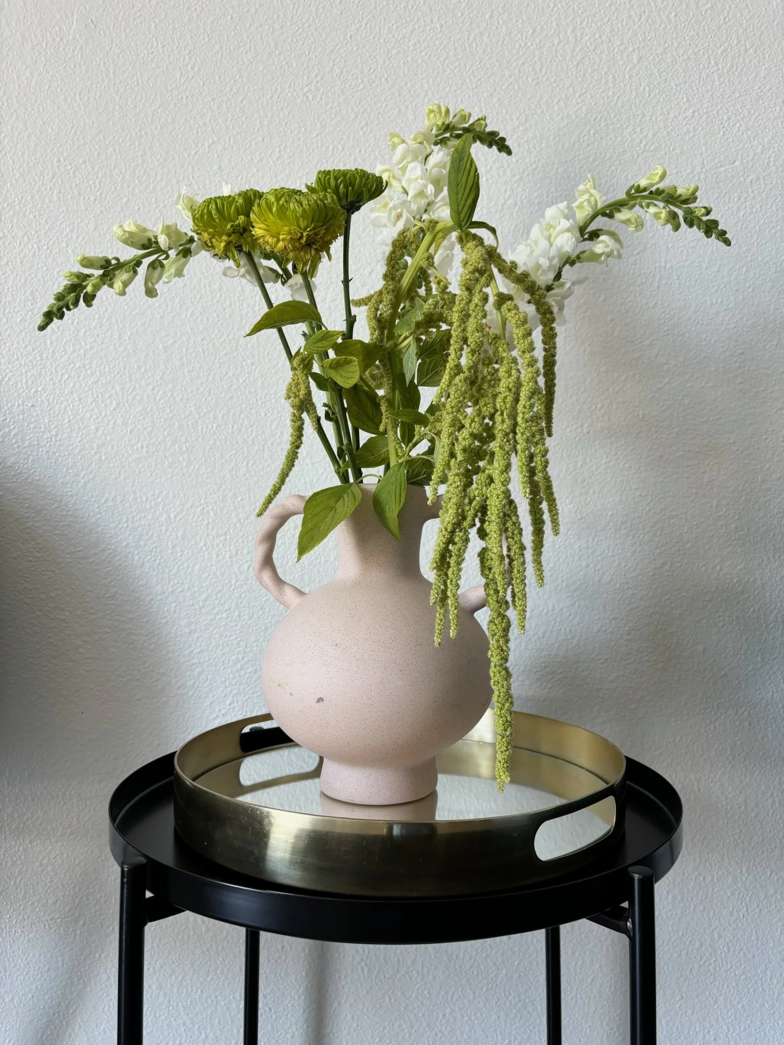 Flower arrangement with white and green flowers in a pink vase on a black and gold tray table against a plain white wall.
