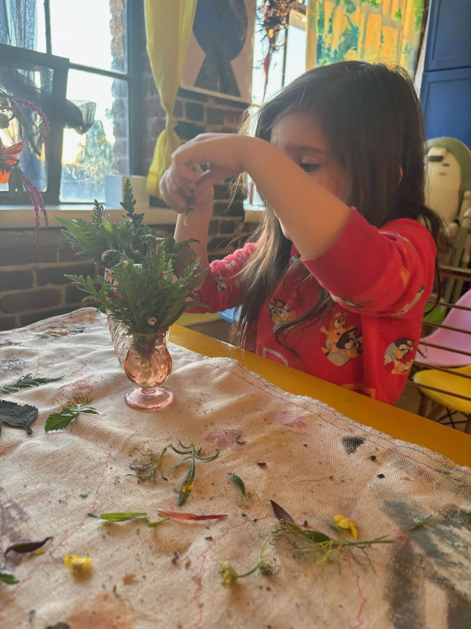 A young girl with long hair wearing a red shirt with cartoon characters, sitting at a yellow table, arranging greenery and flowers in a small pink glass vase, with leaves and flower petals scattered on the table.