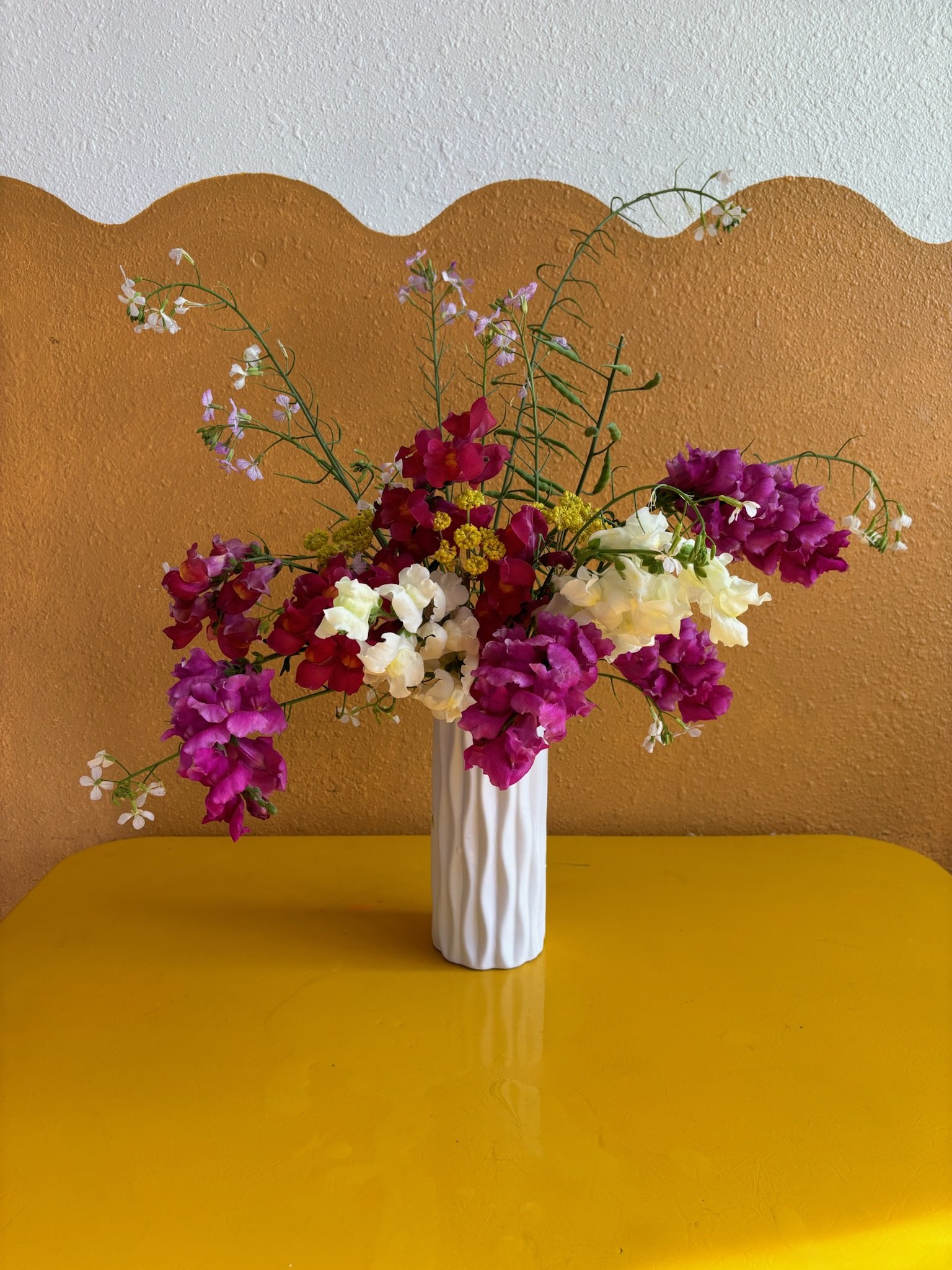 Colorful bouquet of flowers in a white vase on a yellow table, with a two-tone wall background.