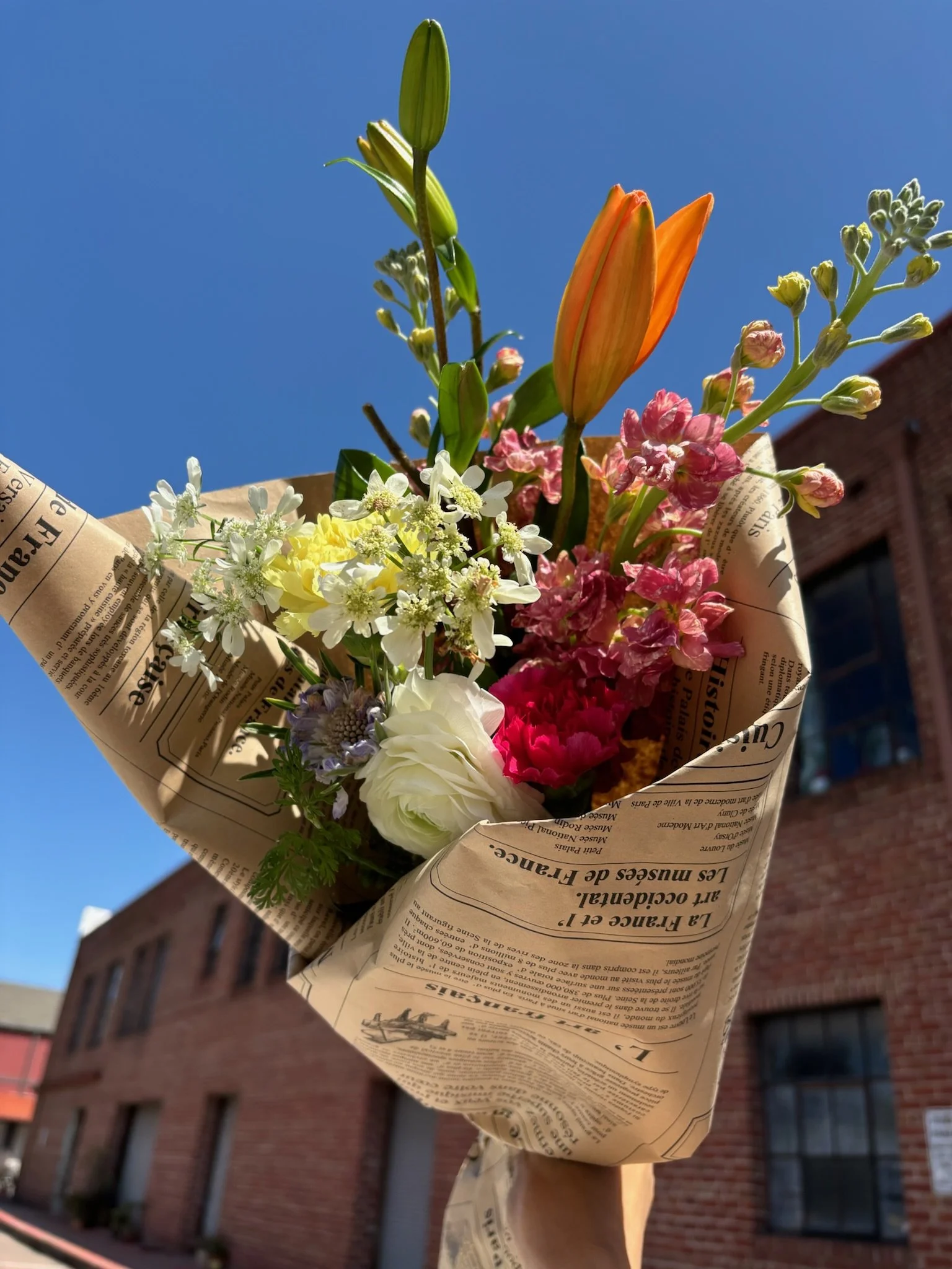 A colorful bouquet of flowers wrapped in newspaper, held up outdoors against a blue sky with brick building in the background.