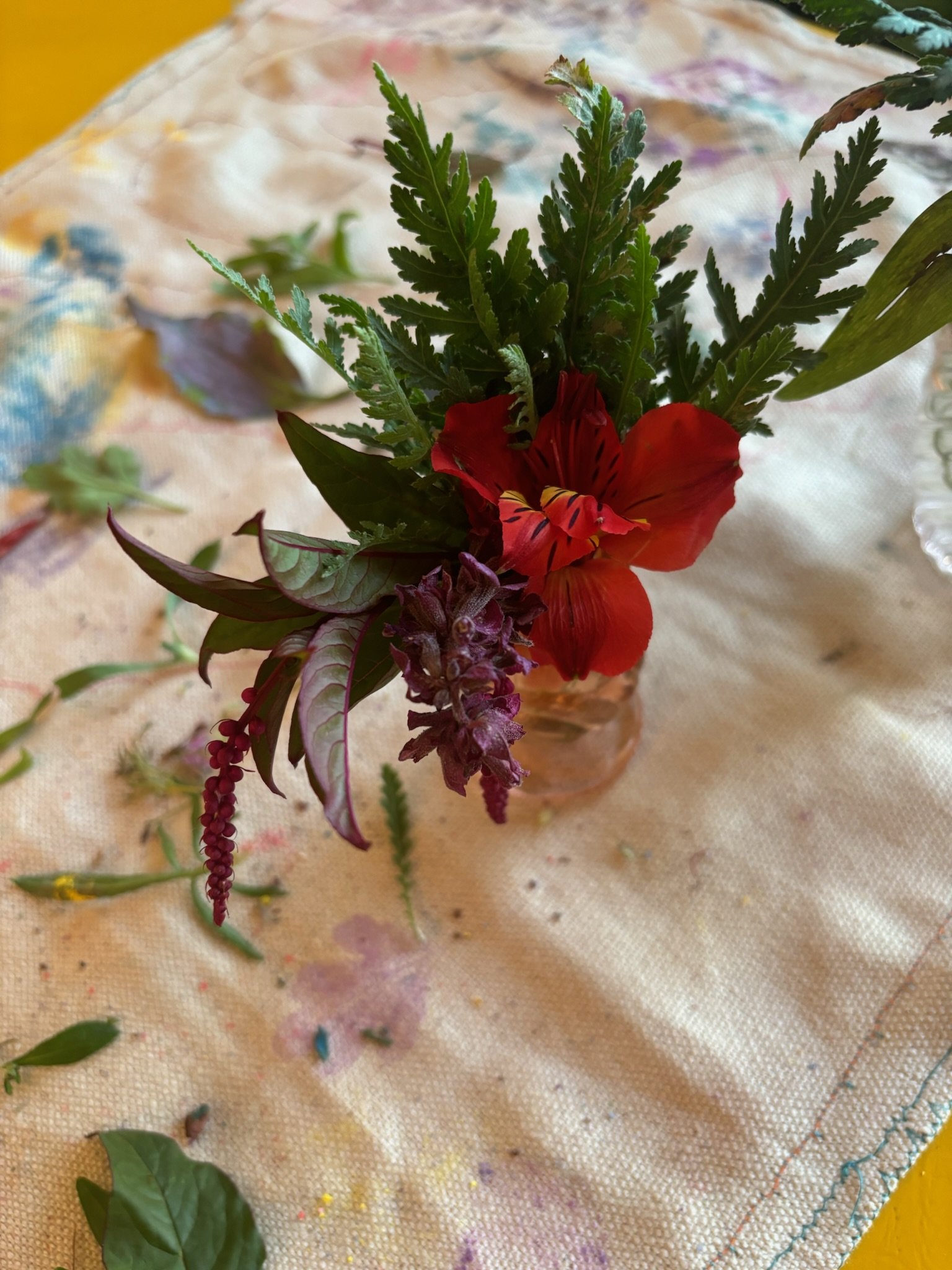 A small flower arrangement in a glass jar with red, purple, and green foliage, placed on a fabric surface with flowers and leaves nearby.