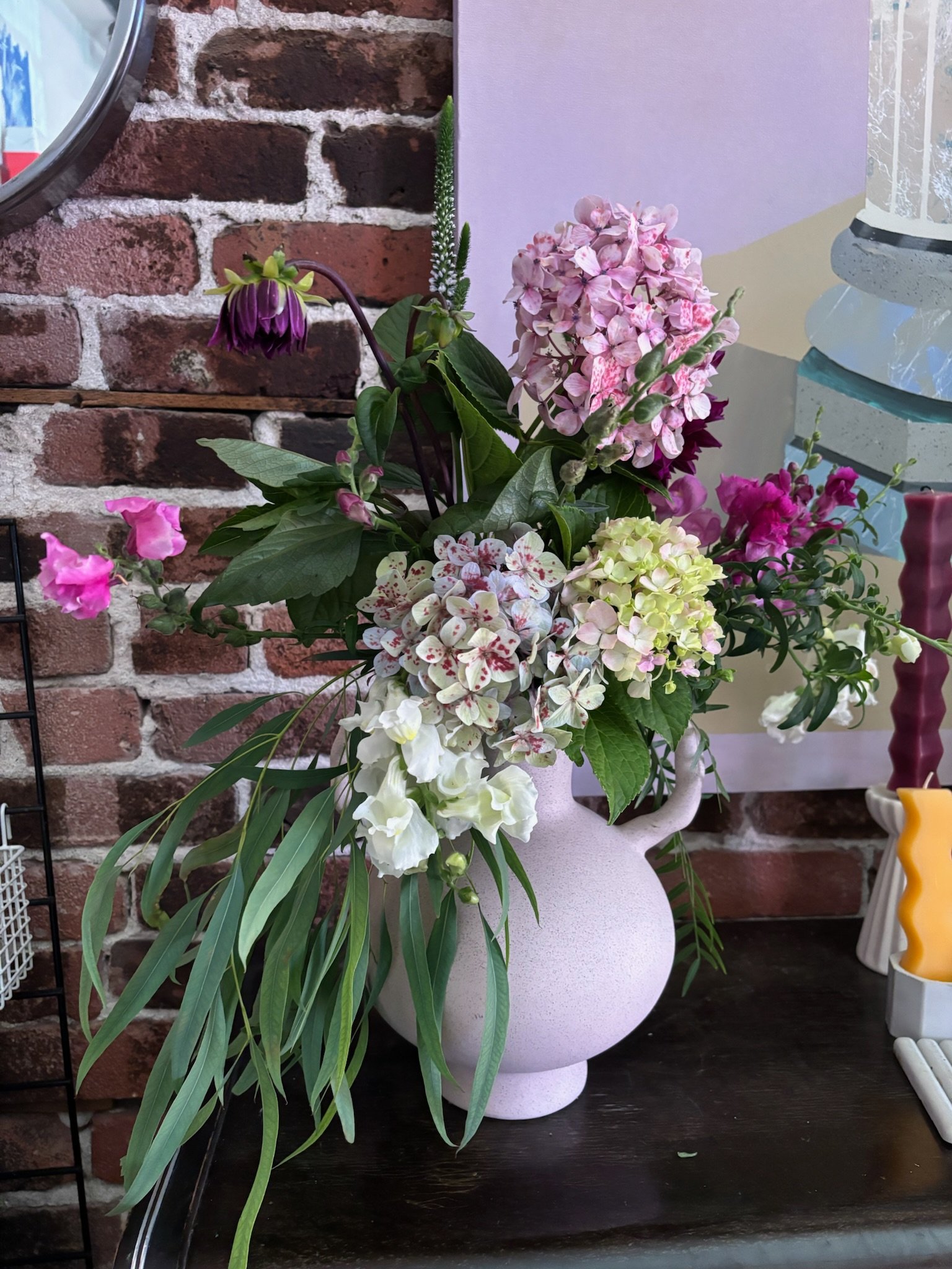 A pink ceramic vase containing an arrangement of mixed flowers, including pink and white hydrangeas, purple and pink flowers, and greenery, placed on a dark table against a brick wall background.