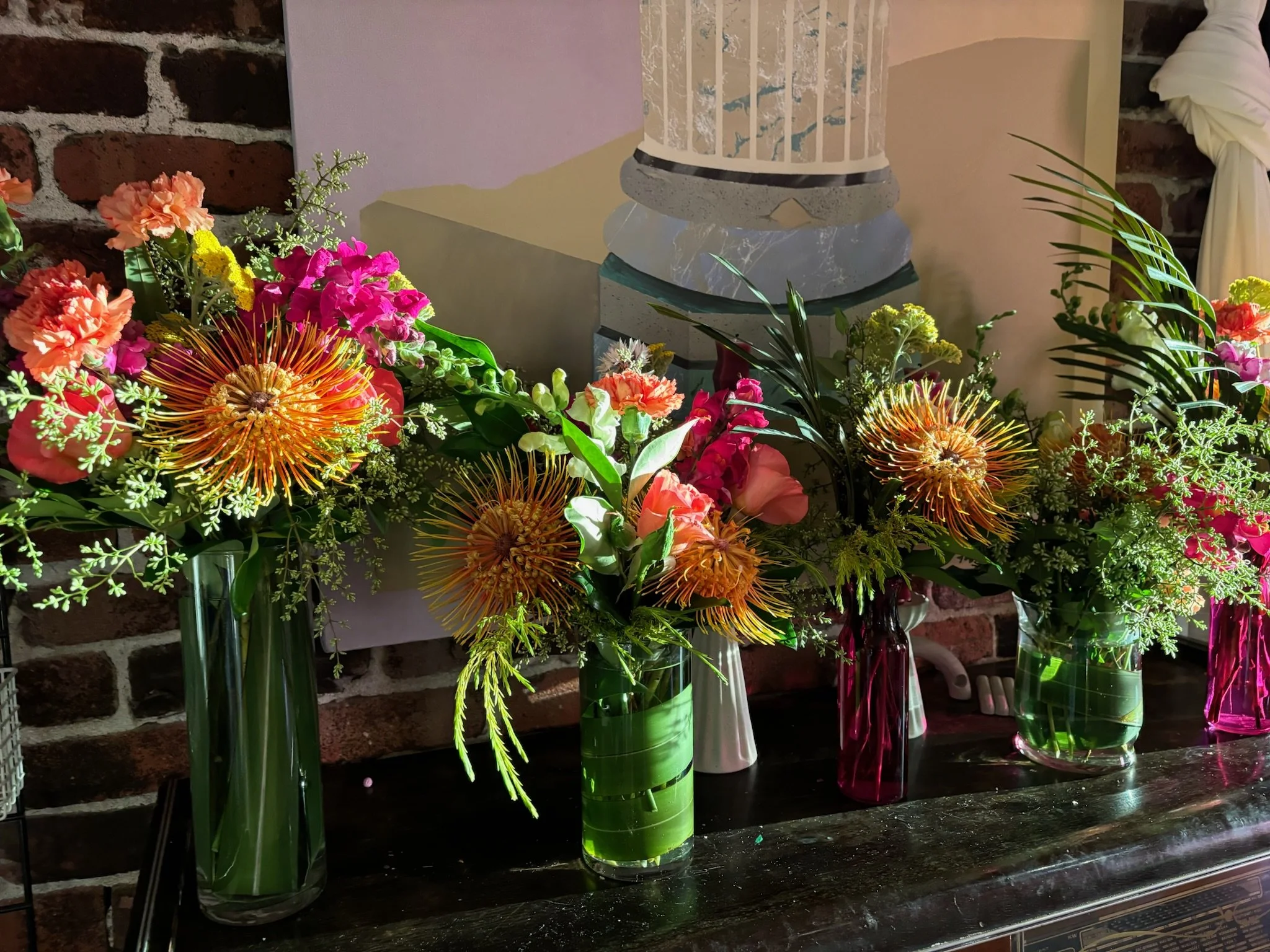 Various colorful flower arrangements in glass vases on a dark wooden table, with a brick wall in the background.