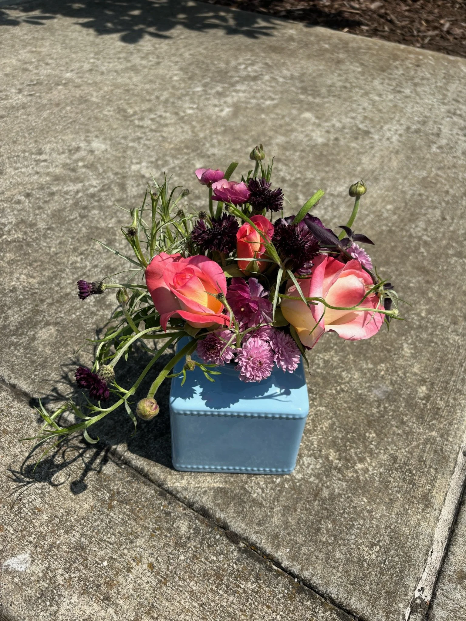 A bouquet of pink and purple flowers in a blue square vase on a concrete surface outdoors in Oakland, CA.