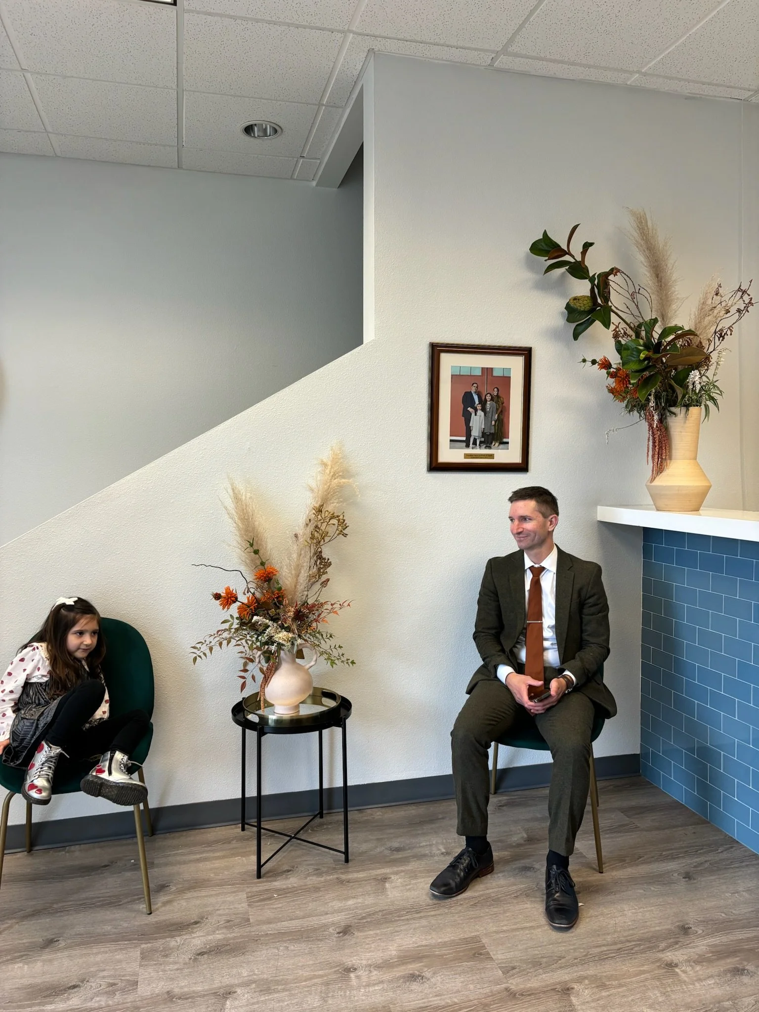 A man in a brown suit and a girl sitting on chairs in a waiting area with floral decorations and a framed photo on the wall.