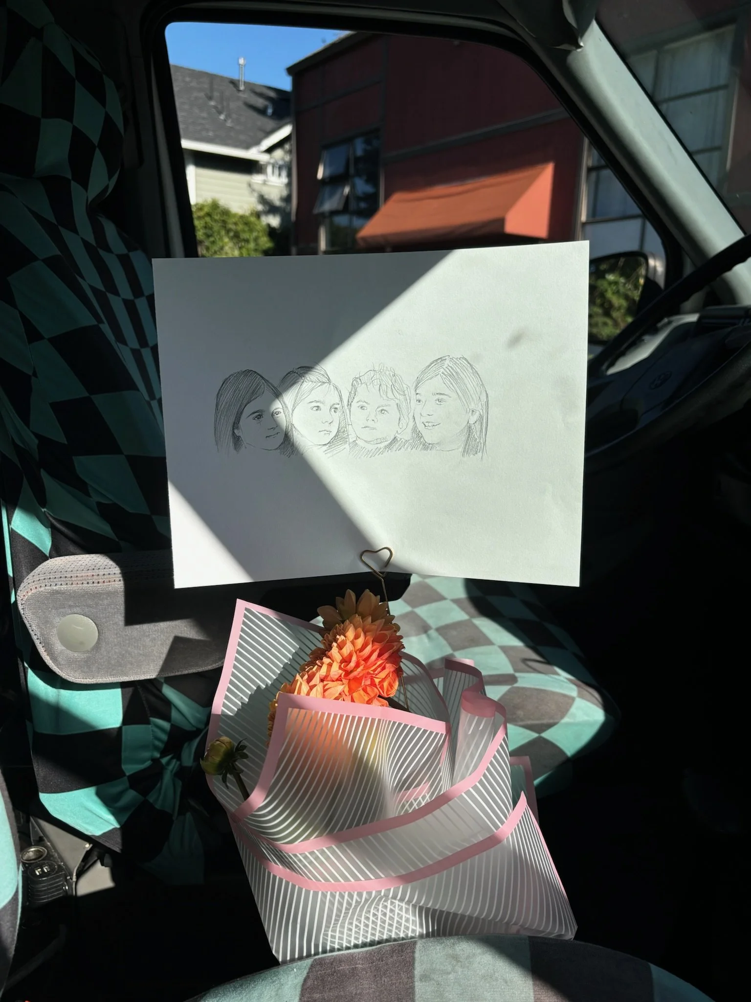 Hand-drawn sketch of four women on a white paper, propped up inside a vehicle, with a gift bag containing flowers in front of it. The vehicle's interior has a patterned fabric, and the outside scene shows houses with blue sky.