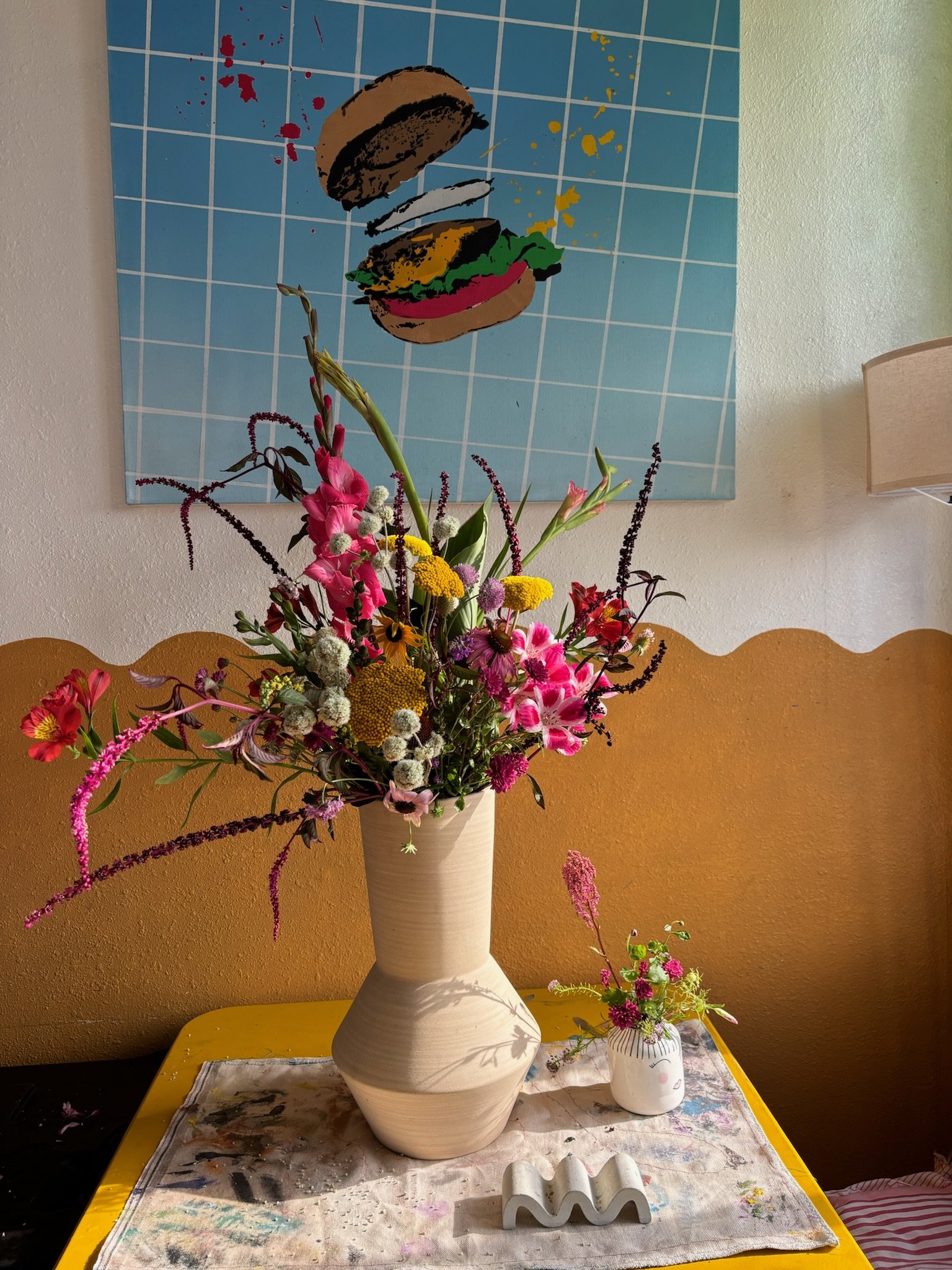 A large beige vase filled with a colorful arrangement of various flowers, including pink lilies, yellow and white blooms, and green foliage, on a table with a smaller white flower vase and decorative object nearby. A pastel-colored wall and a blue wa