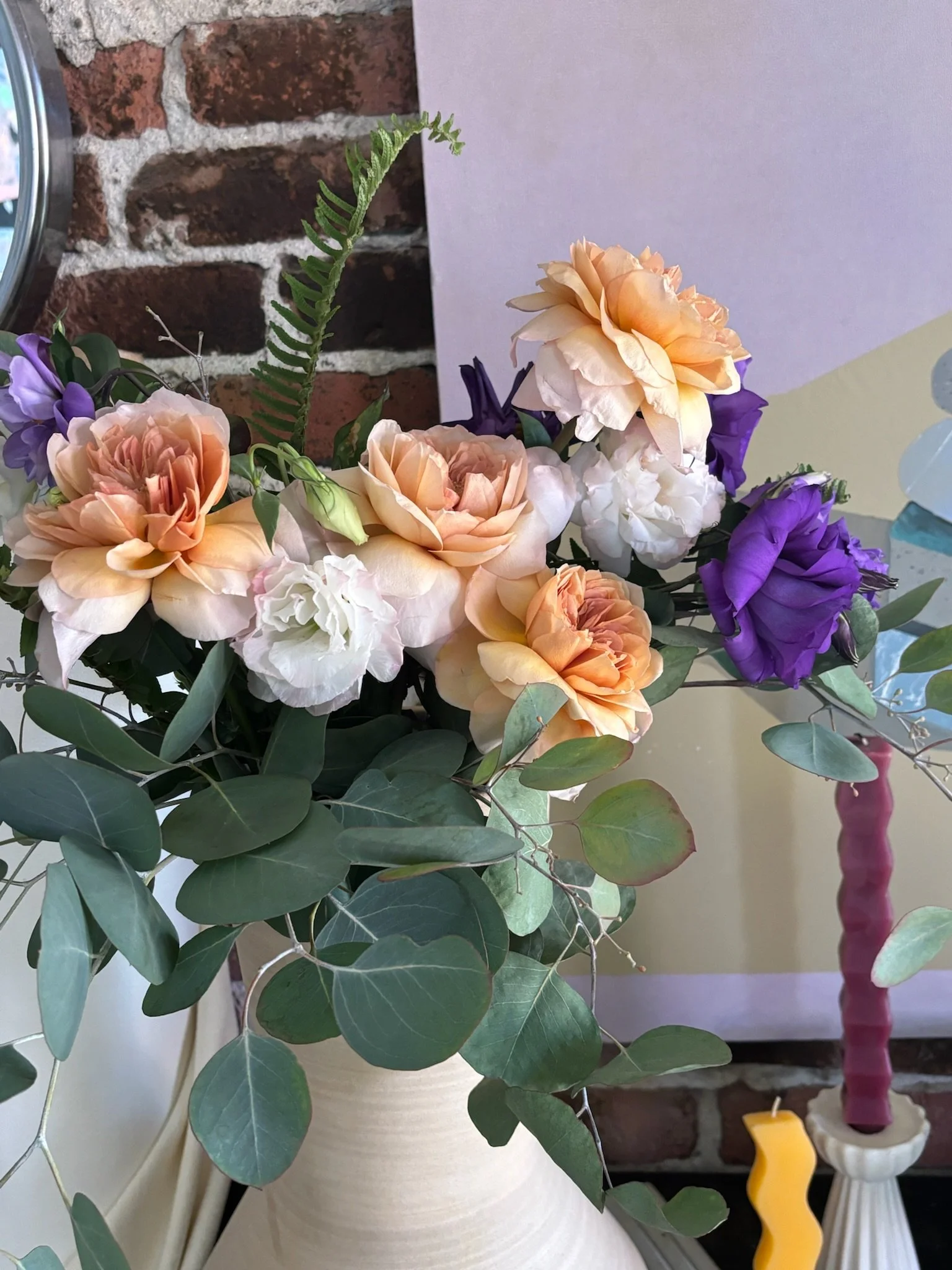 A bouquet of peach and white roses, purple lisianthus, eucalyptus leaves, and a fern in a white ceramic vase.