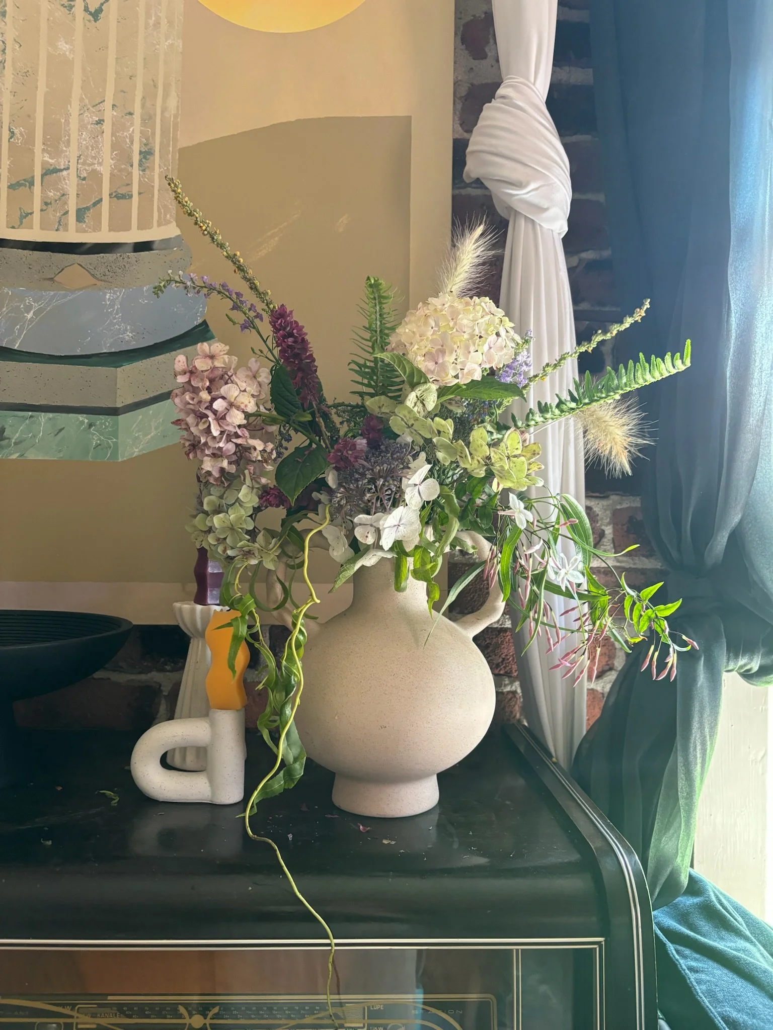 A beige ceramic vase with a wide body and narrow neck holding a colorful bouquet of hydrangeas, ferns, and other flowers. The vase is placed on a dark surface next to a white ceramic sculpture.