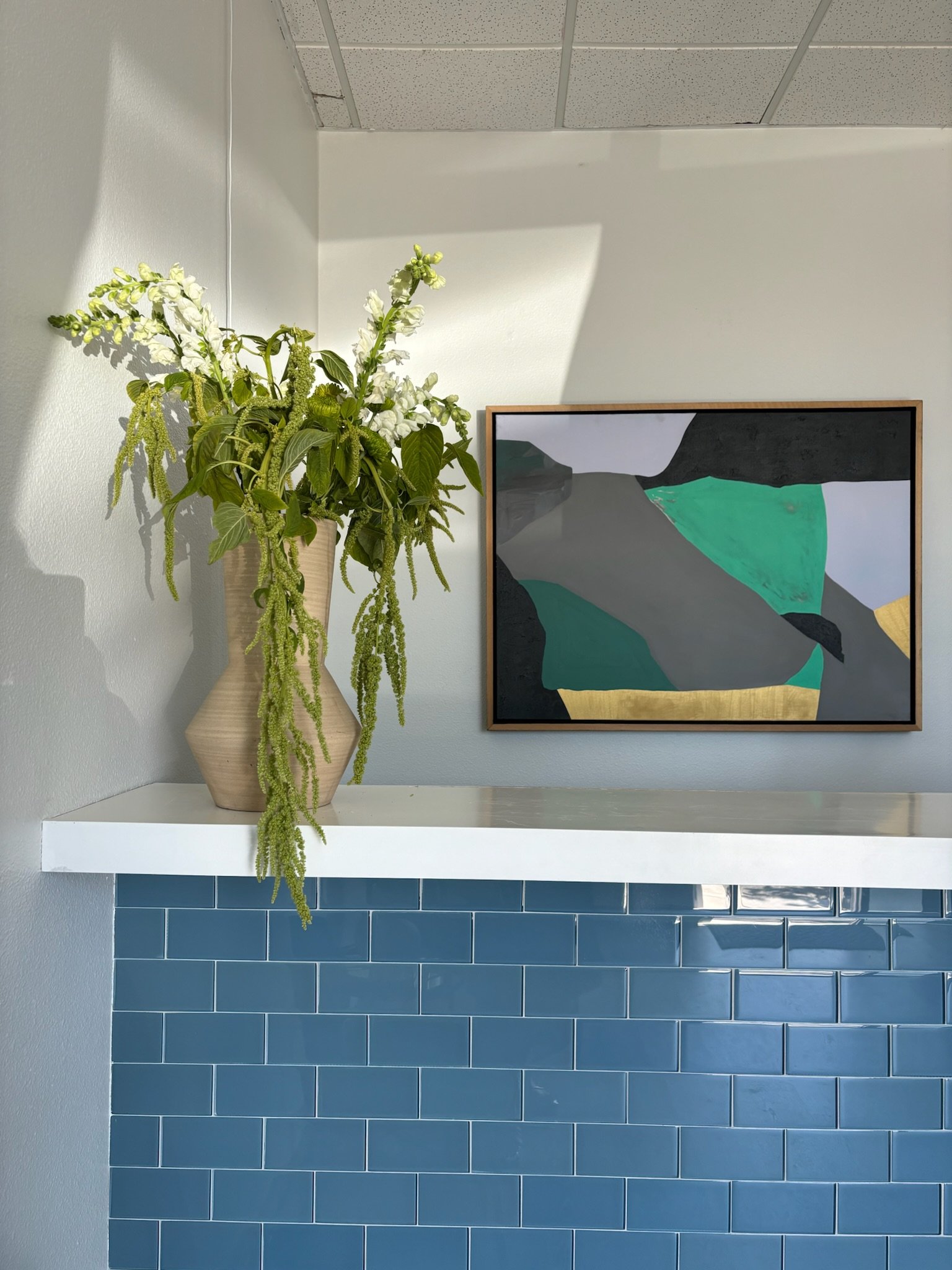 A beige vase with white flowers and green foliage on a white ledge, next to a framed abstract landscape painting on a light gray wall, with blue tiles below.