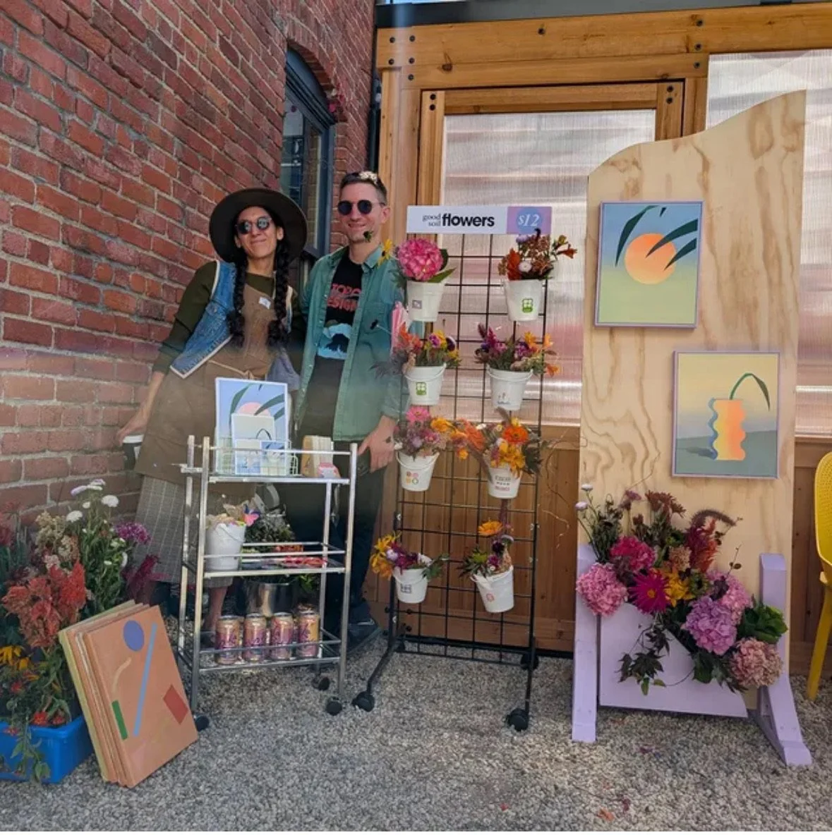 A man and woman stand at a flower stall with various colorful flowers, artwork, and informational signs, set against a brick wall and wooden display panels.