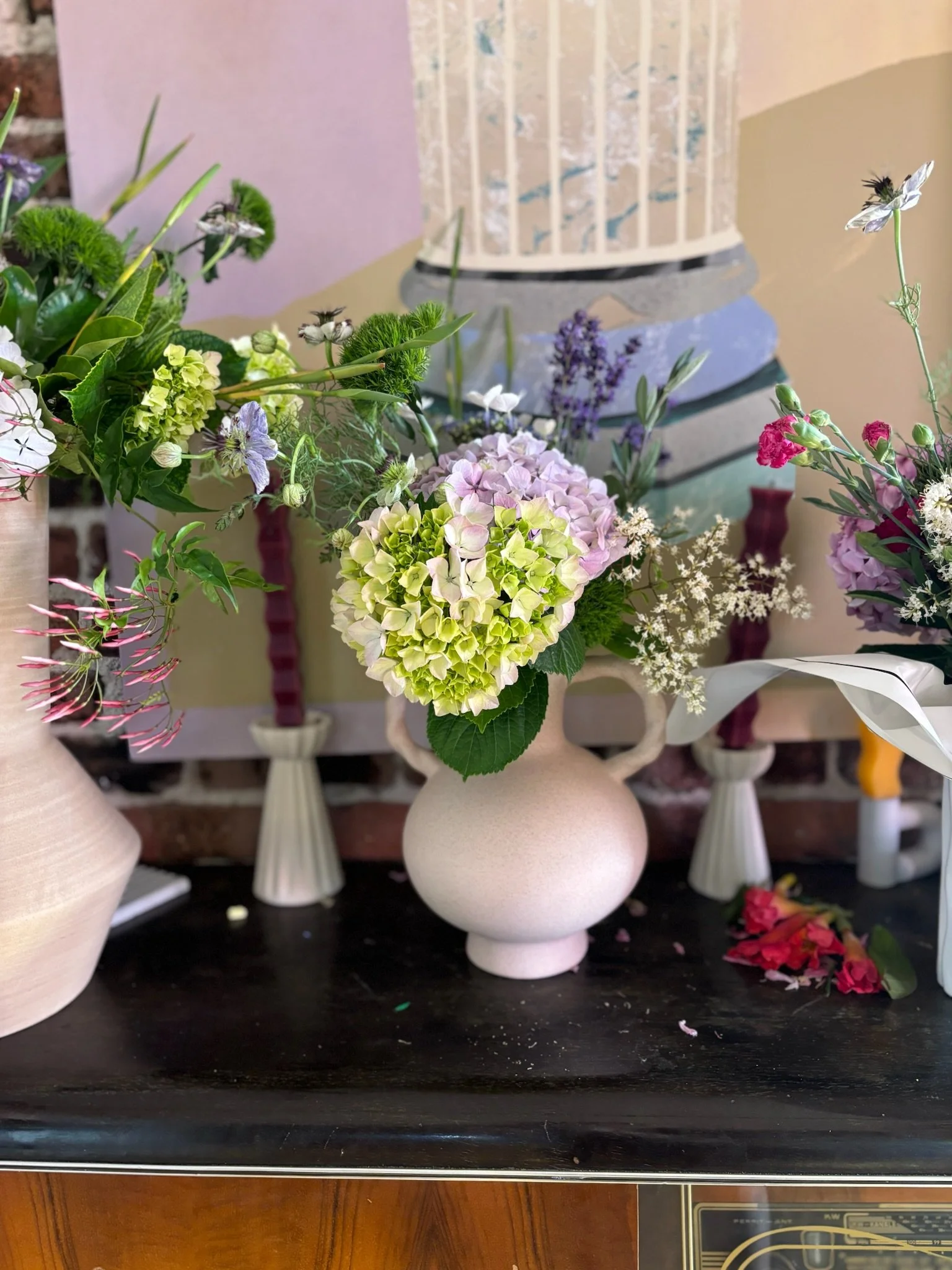 A white vase filled with pink, lavender, and green hydrangea flowers is displayed on a black table, with other floral arrangements and a painting in the background.