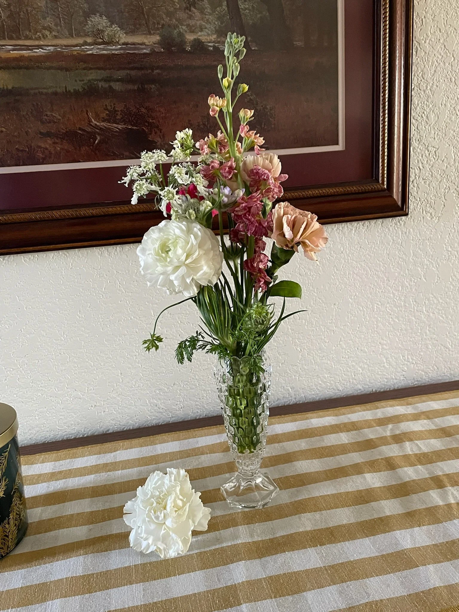 A glass vase with a bouquet of mixed flowers including white, pink, and mauve blossoms, placed on a table with a striped tablecloth; a white flower petal is on the table beside the vase.