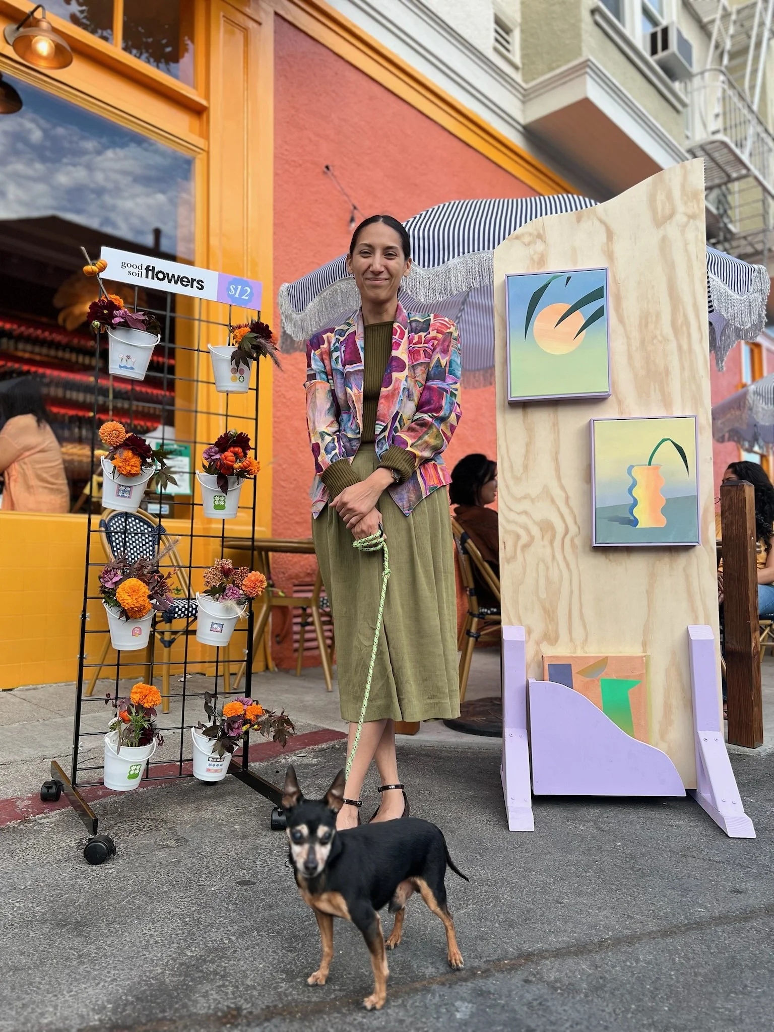 Woman standing outside a flower shop with a dog on a leash, colorful artwork displayed on a wooden board, and a pink and orange building in the background.